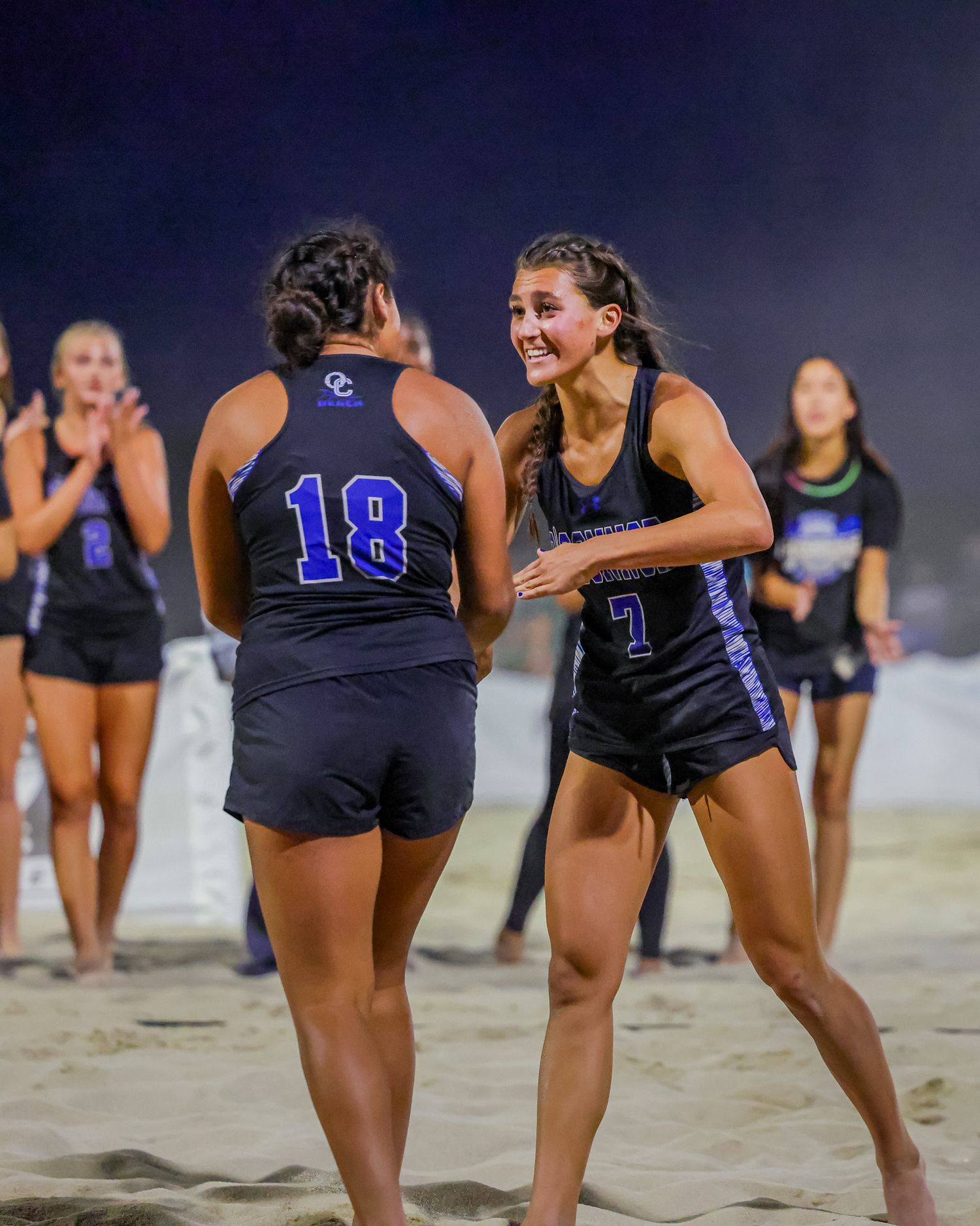 Two female beach volleyball players in dark uniforms celebrating together on a sandy court at night.