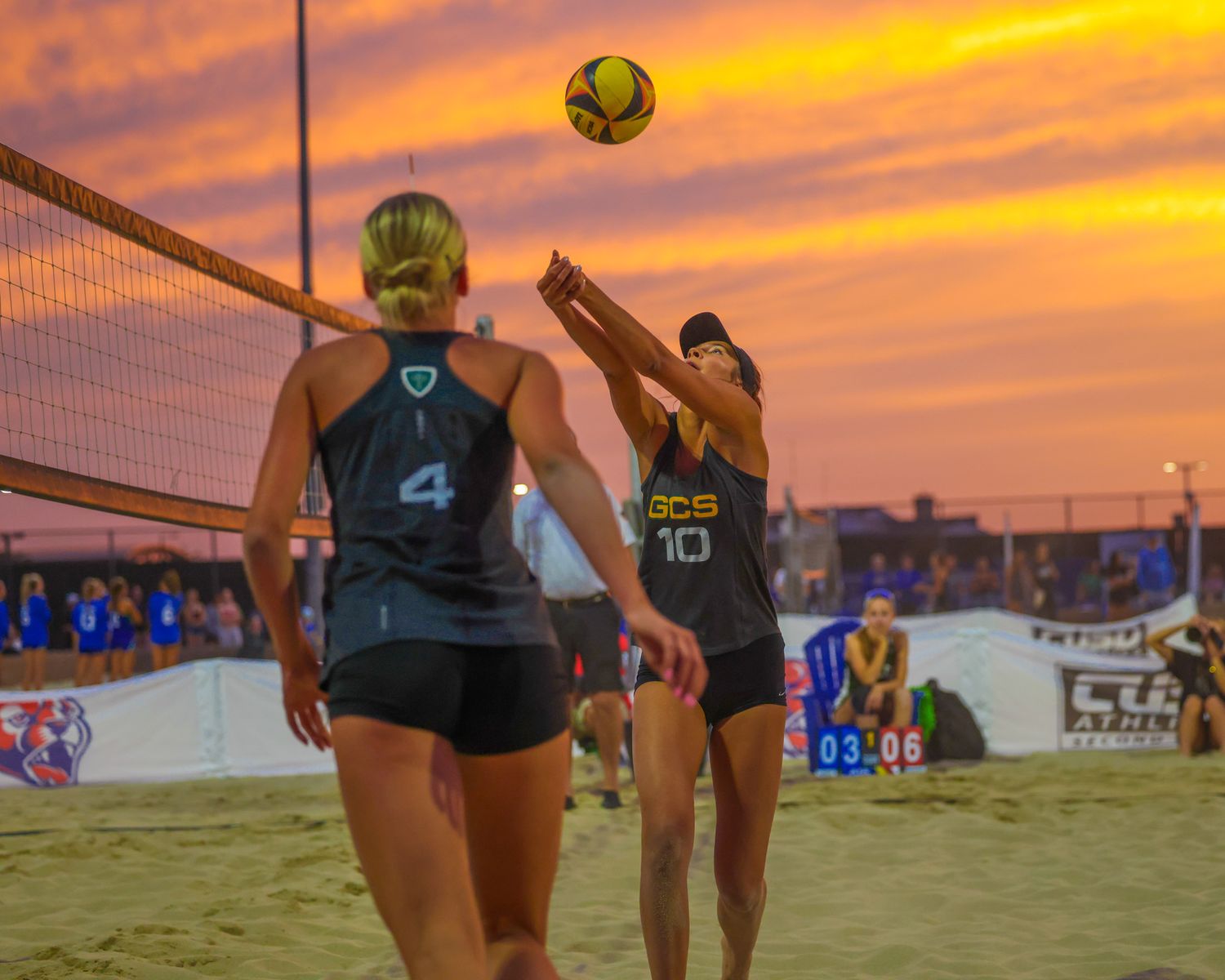 Two female beach volleyball players at the net during a competitive sunset match on a sand court.