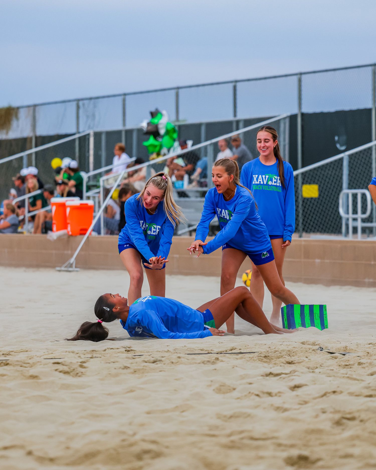 Xavier beach volleyball teammates in blue uniforms helping fallen player off sandy court while laughing together.
