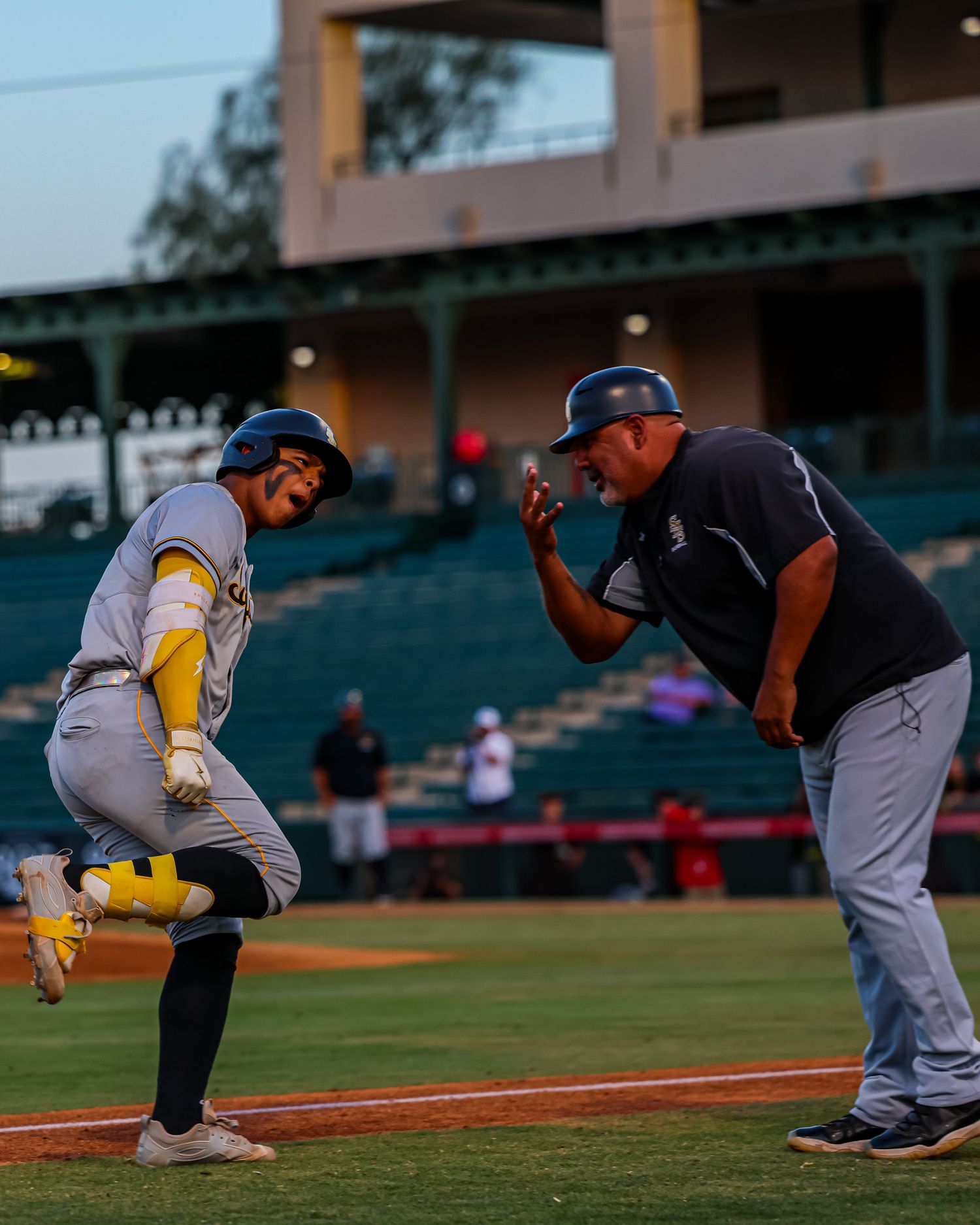 Baseball coach celebrates with player rounding third base during sunny game.