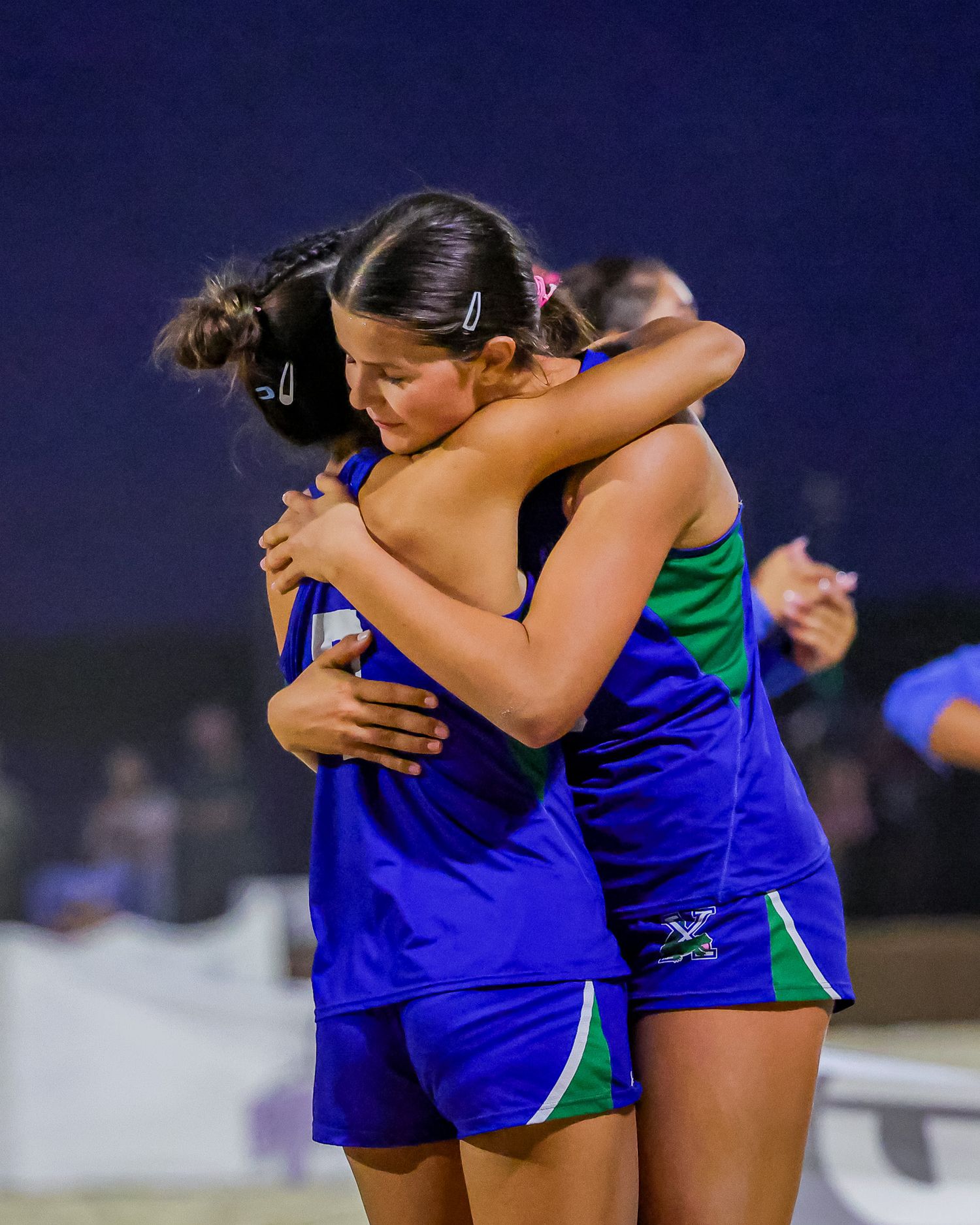 Two female beach volleyball players in blue and green uniforms sharing an emotional hug after a match.