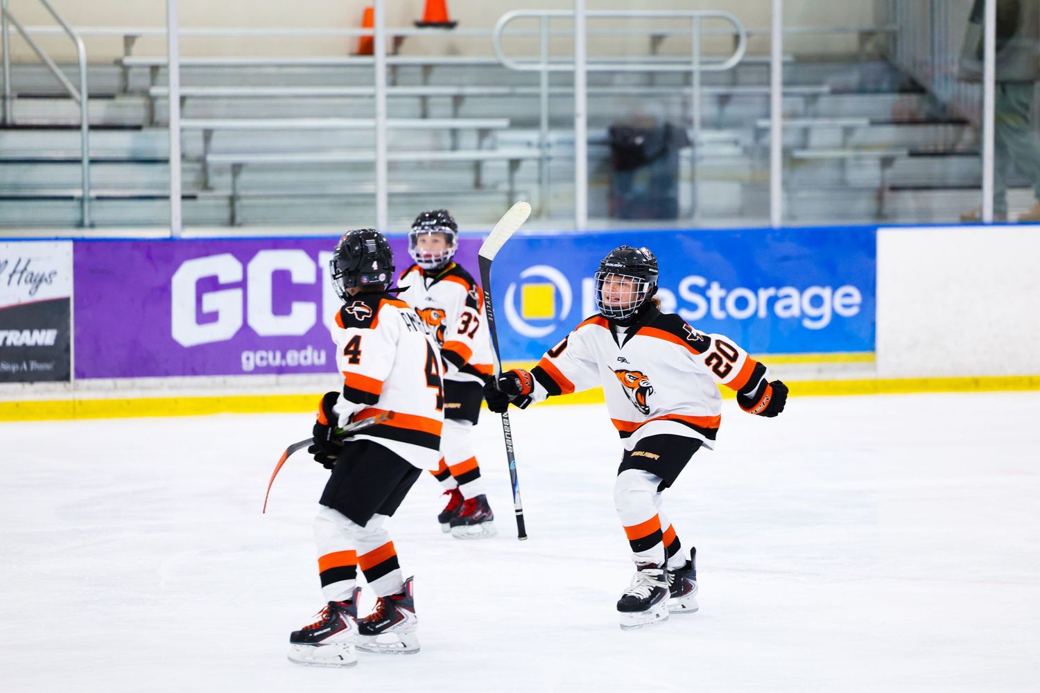 Young hockey players in black and orange uniforms skate on indoor ice rink during a youth hockey game.