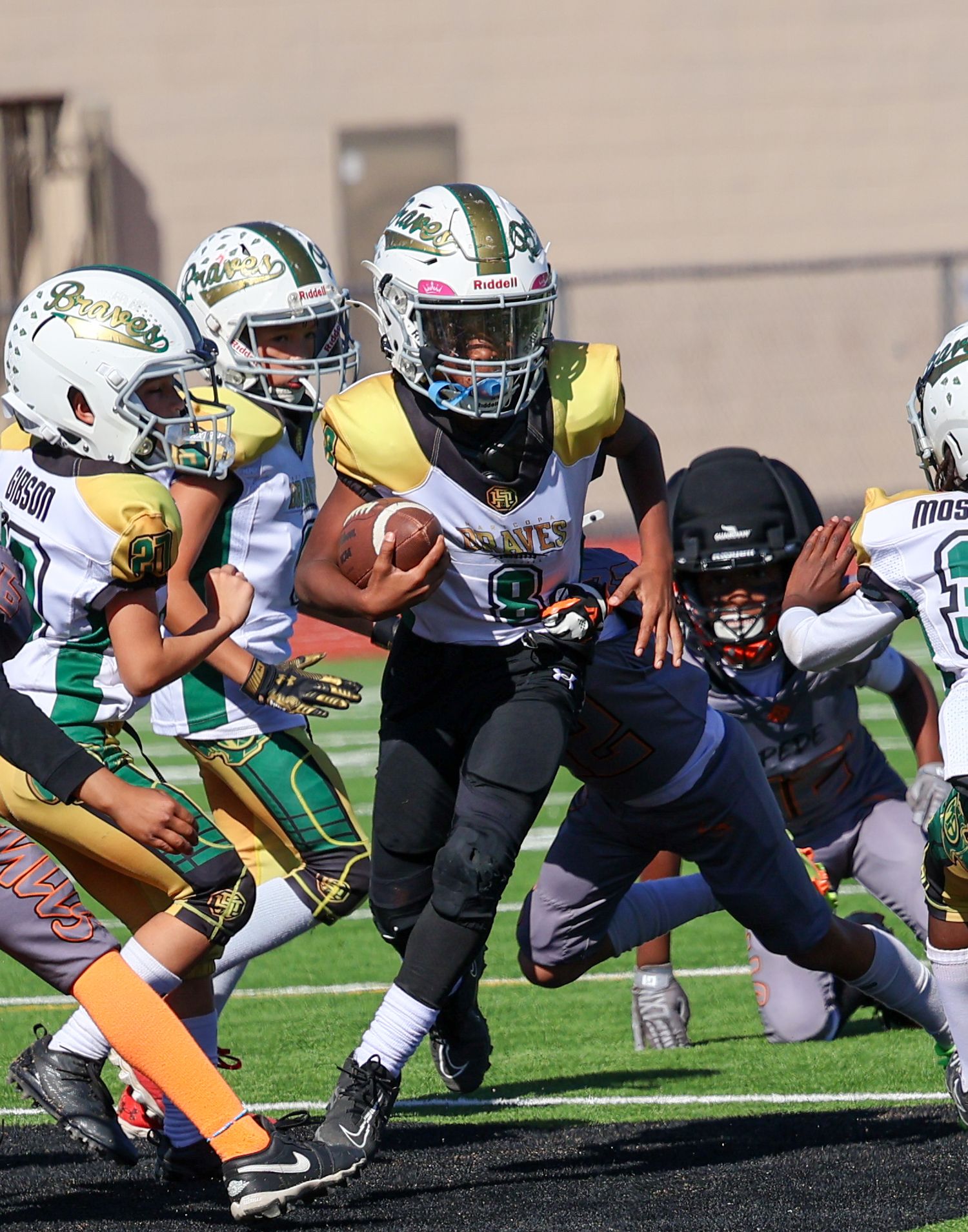 Youth football player in green jersey runs with ball through defenders during game.