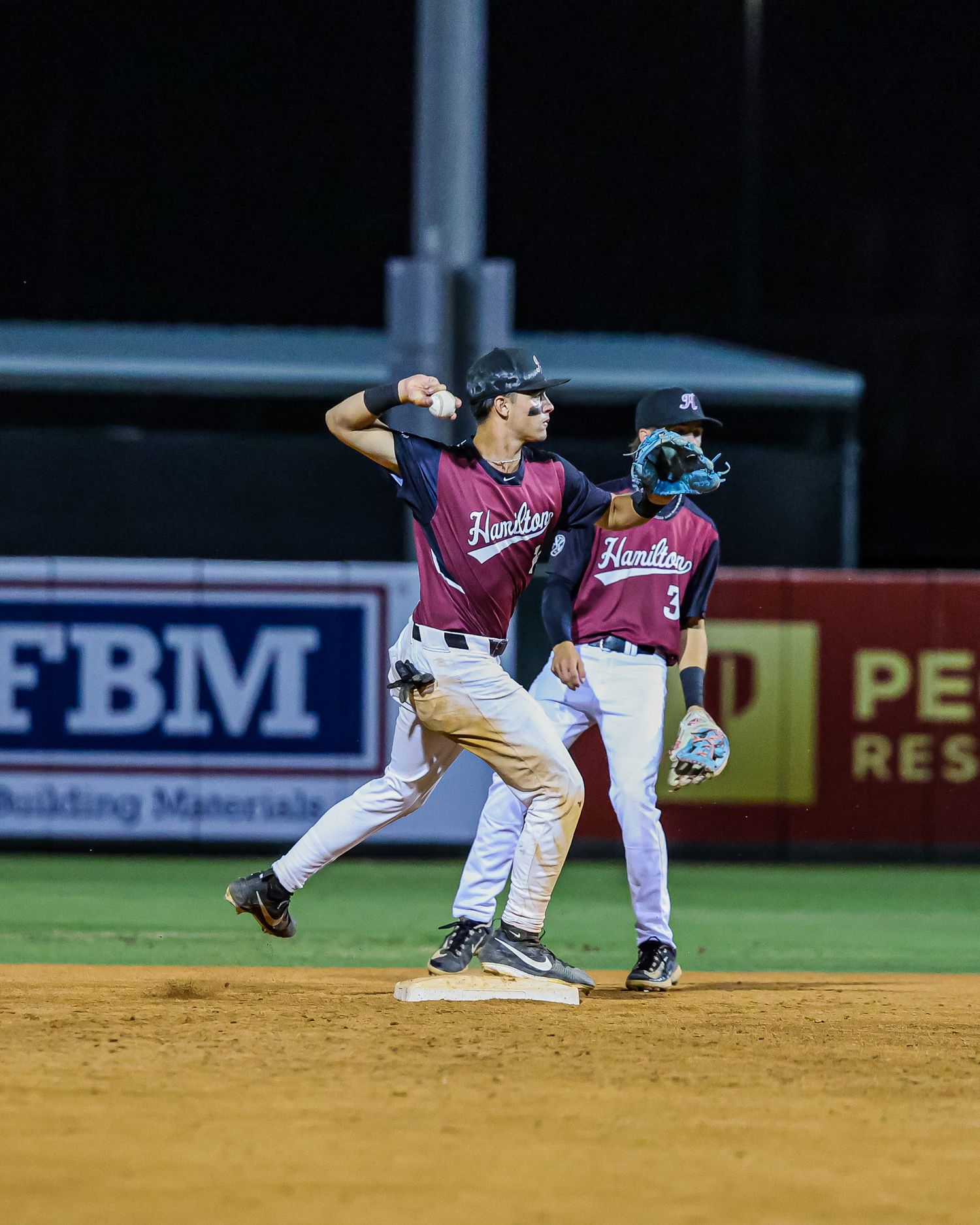 Hamilton baseball infielder throws to first base during a night game with teammate watching.