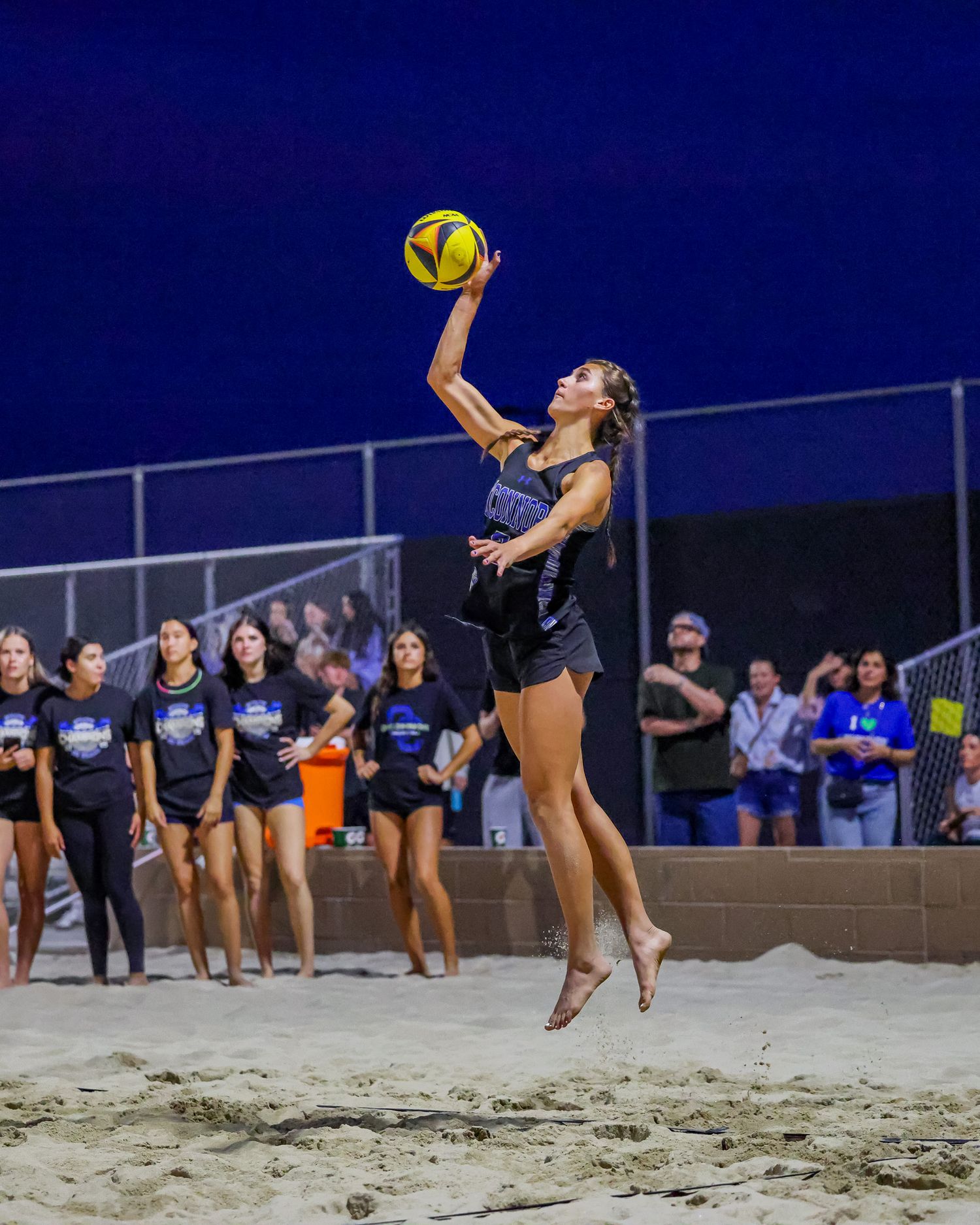 Female beach volleyball player leaping to set the ball during a night game with spectators watching.