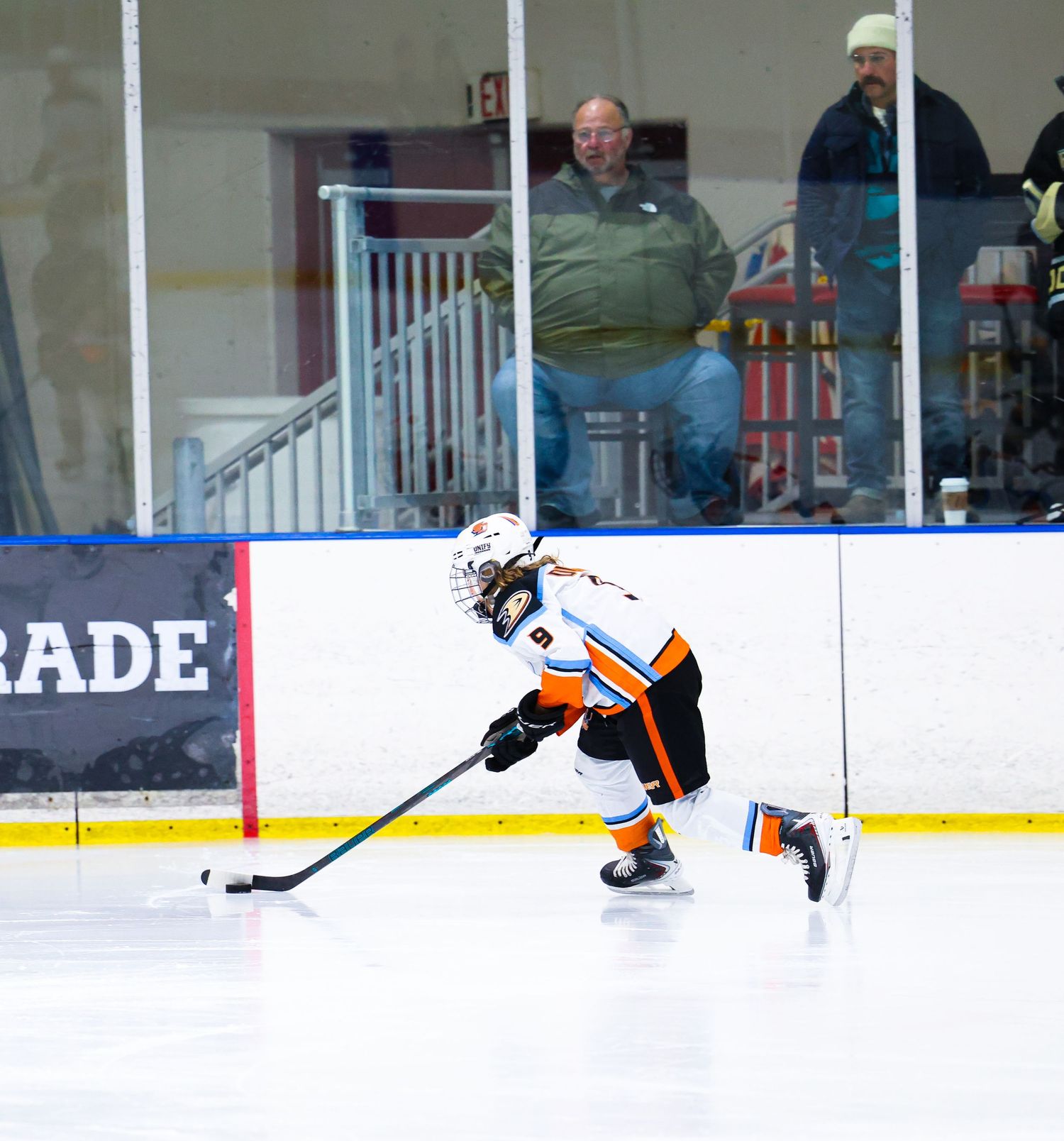 Ice hockey player in orange and white uniform skating with the puck along the boards.