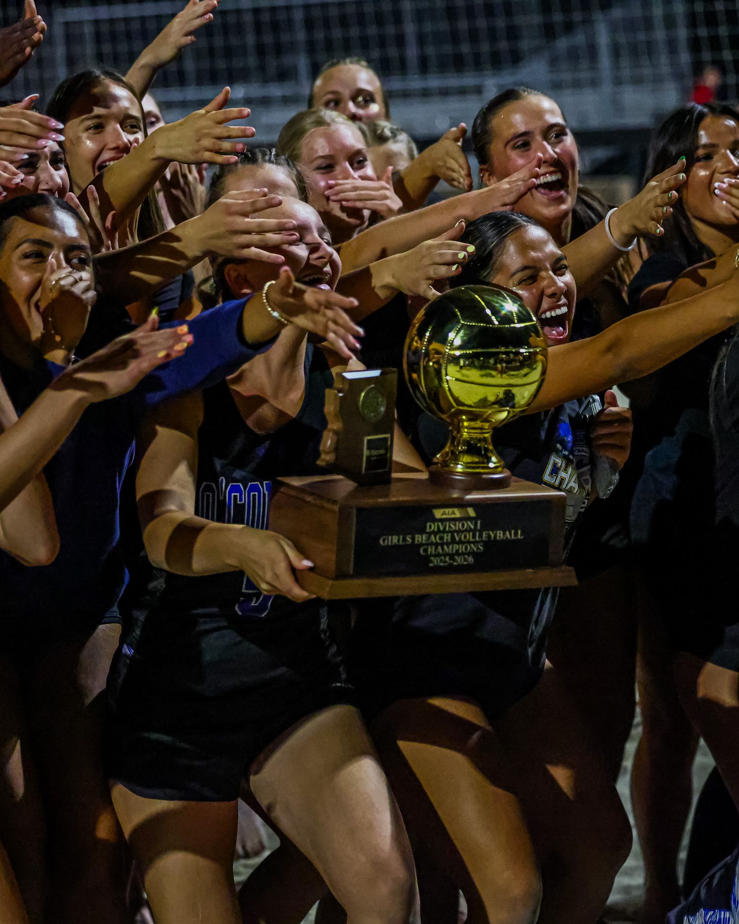 Girls beach volleyball team celebrates winning Division I championship trophy on outdoor court at night.