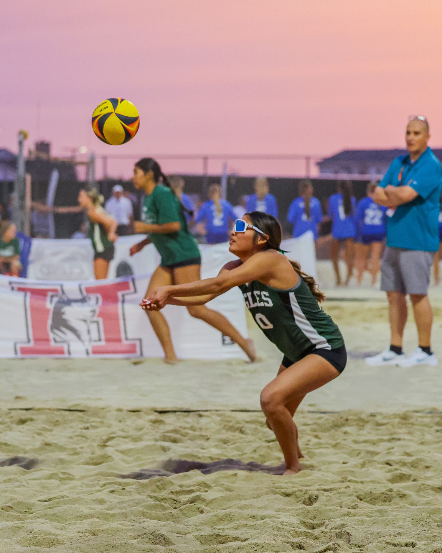 Female beach volleyball player in green jersey digging a yellow ball during a sunset tournament on sand court.
