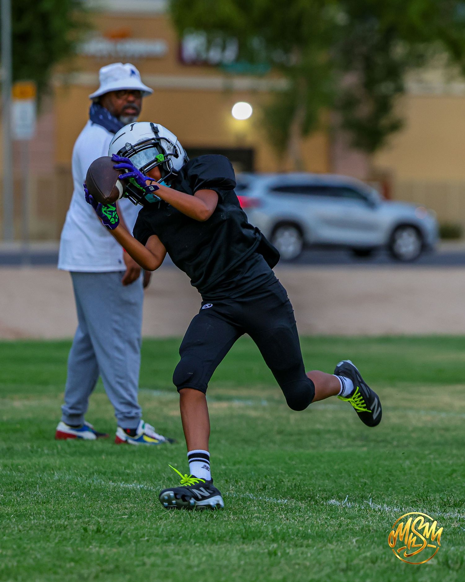 Youth football player catches pass during evening practice with coach watching nearby.