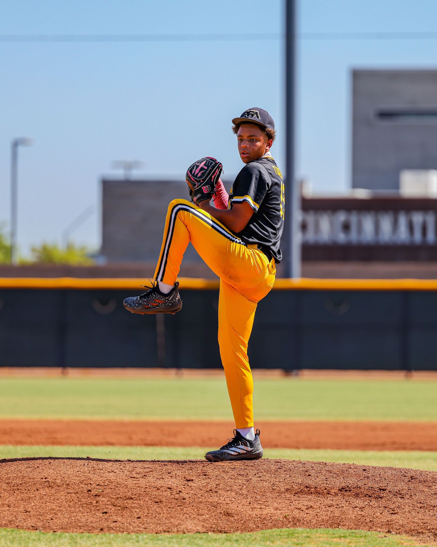 Youth baseball pitcher in gold and black uniform winding up on pitching mound.