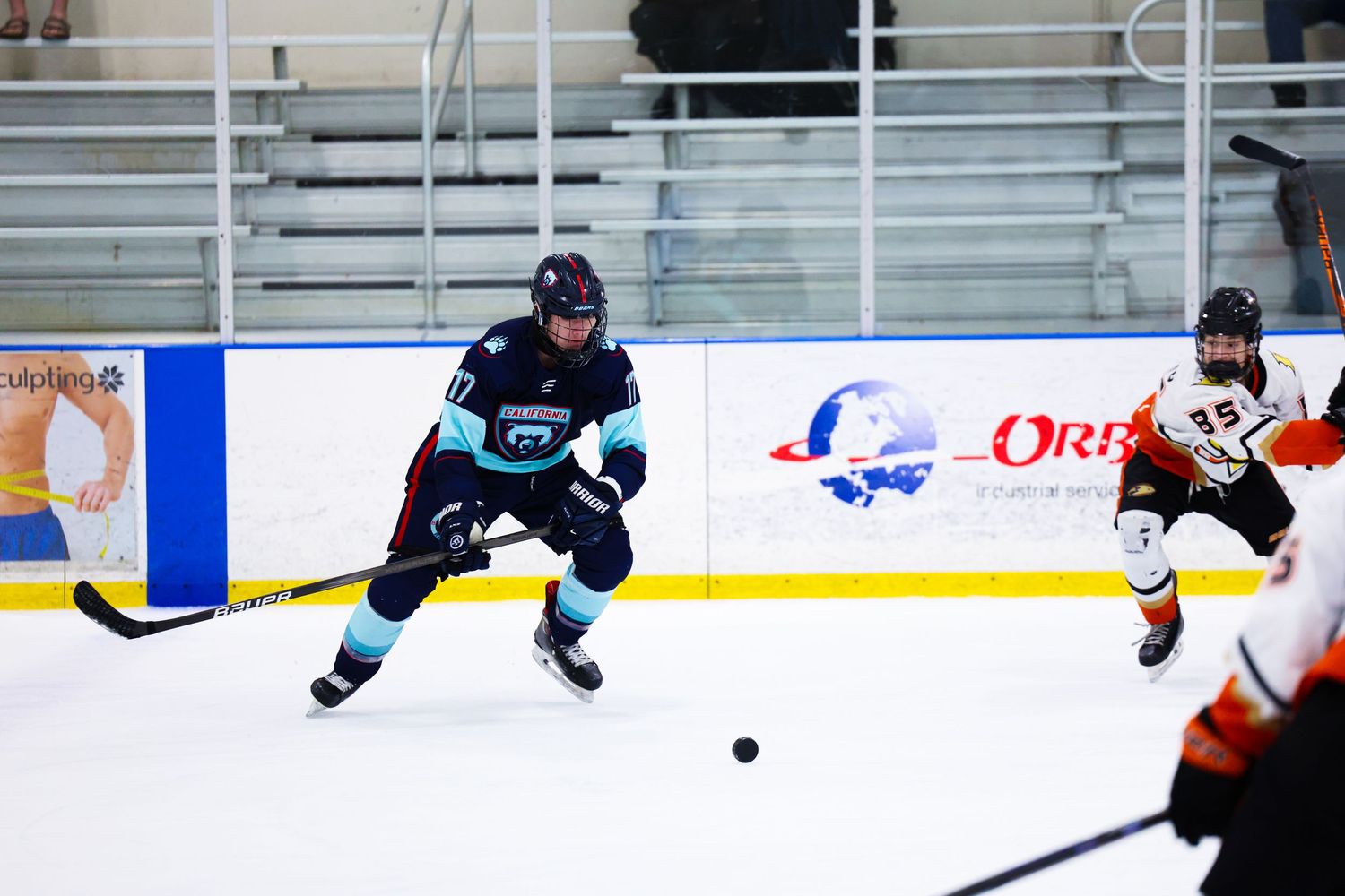 Ice hockey player in teal uniform controls the puck while being pursued by opponent in orange jersey.