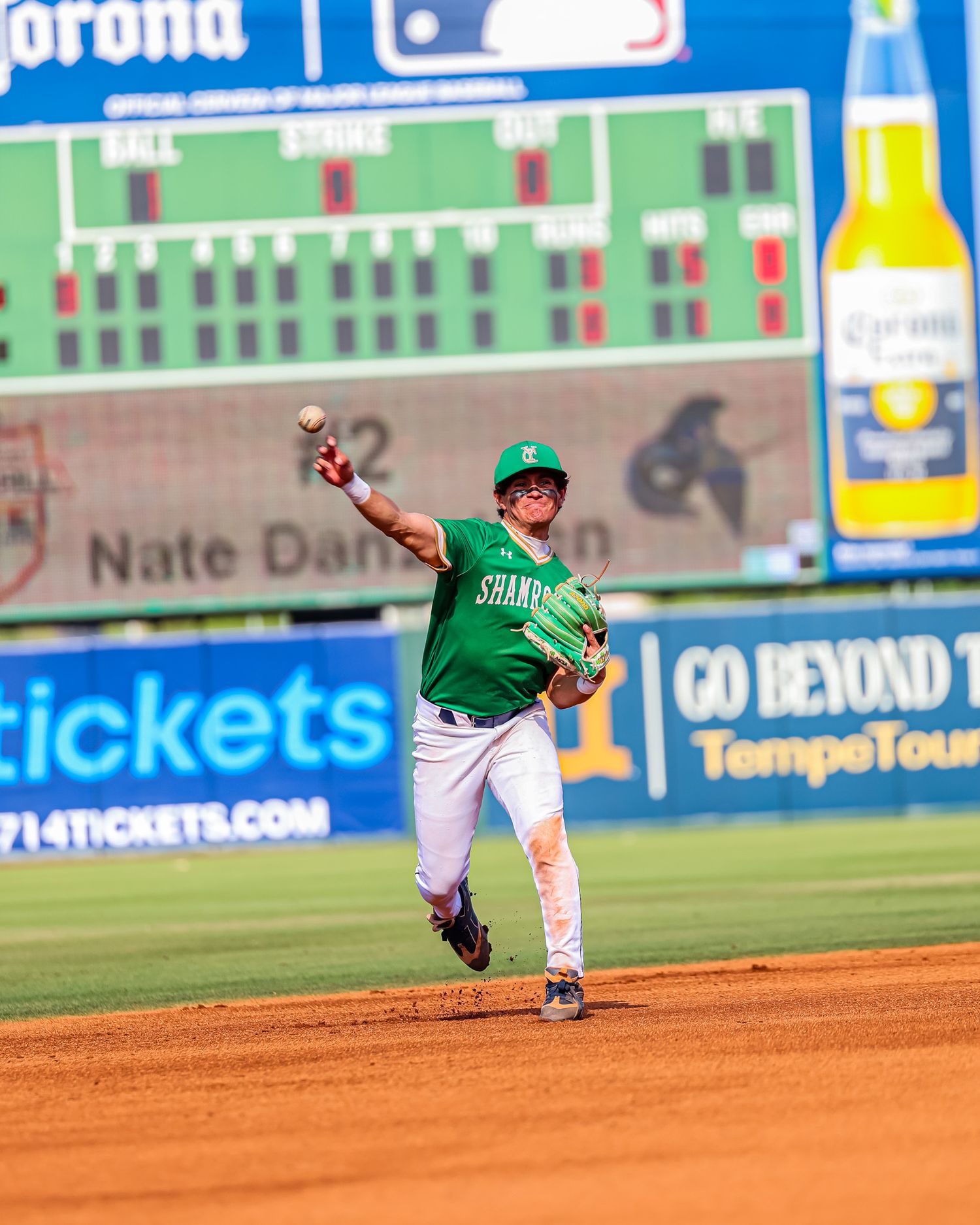 Shamrock baseball player fields and throws from second base in front of a scoreboard.