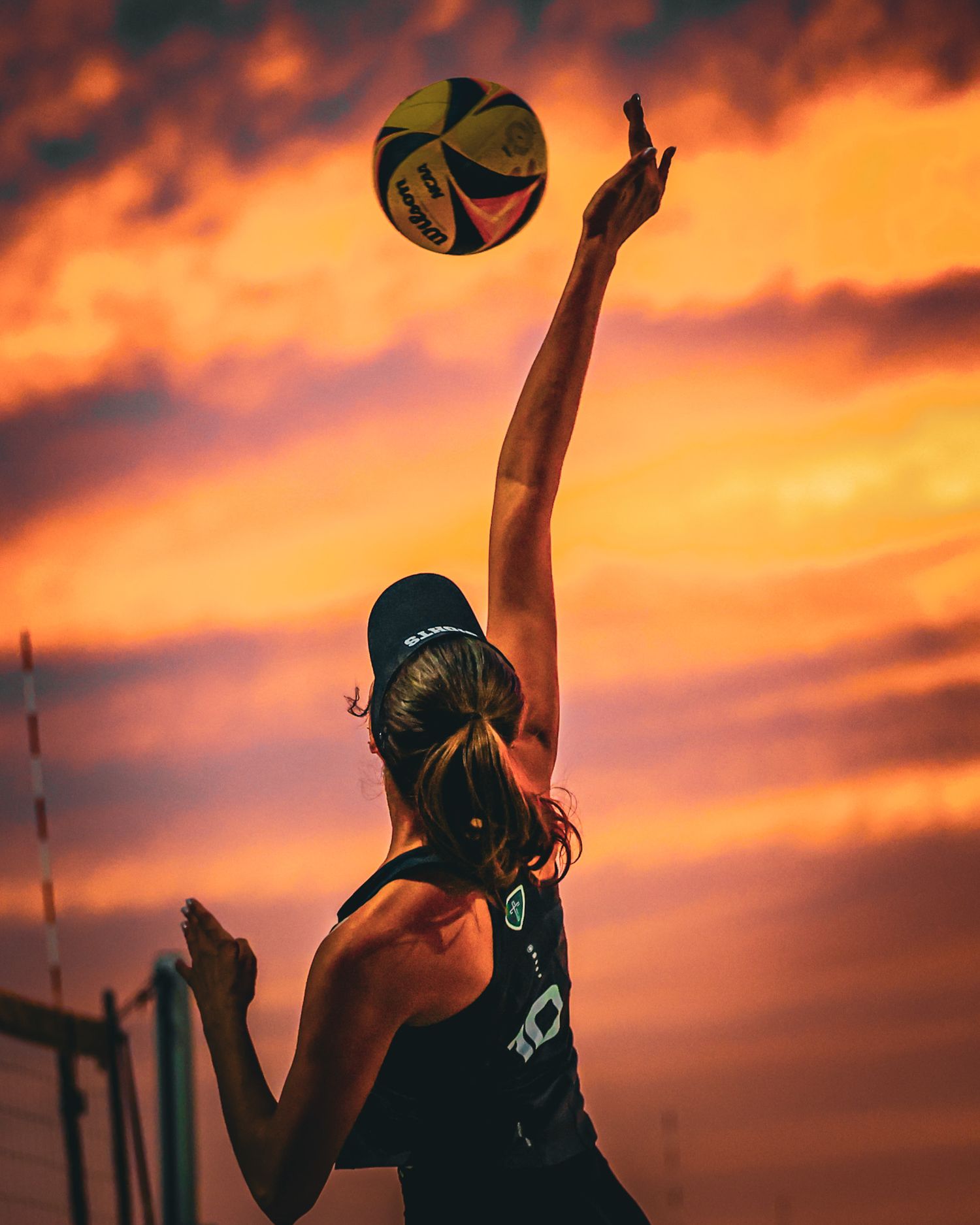 Female beach volleyball player jumping to spike ball against dramatic orange sunset sky during outdoor match.