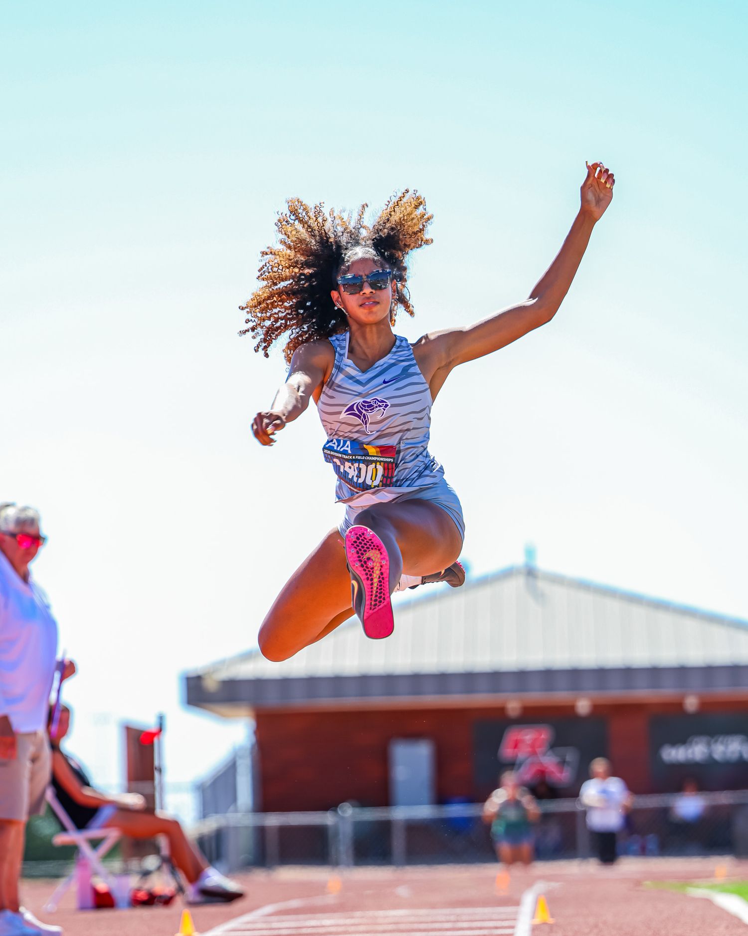 Female track athlete performs long jump mid-air under bright blue sky.