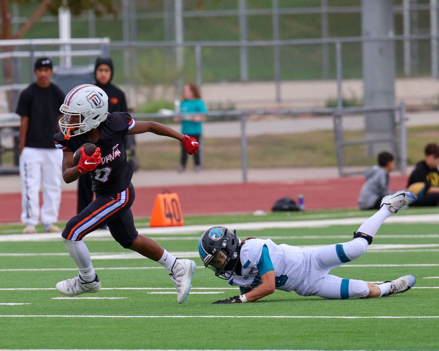 A football player in red and black uniform evades a diving tackler in teal during a high school game.