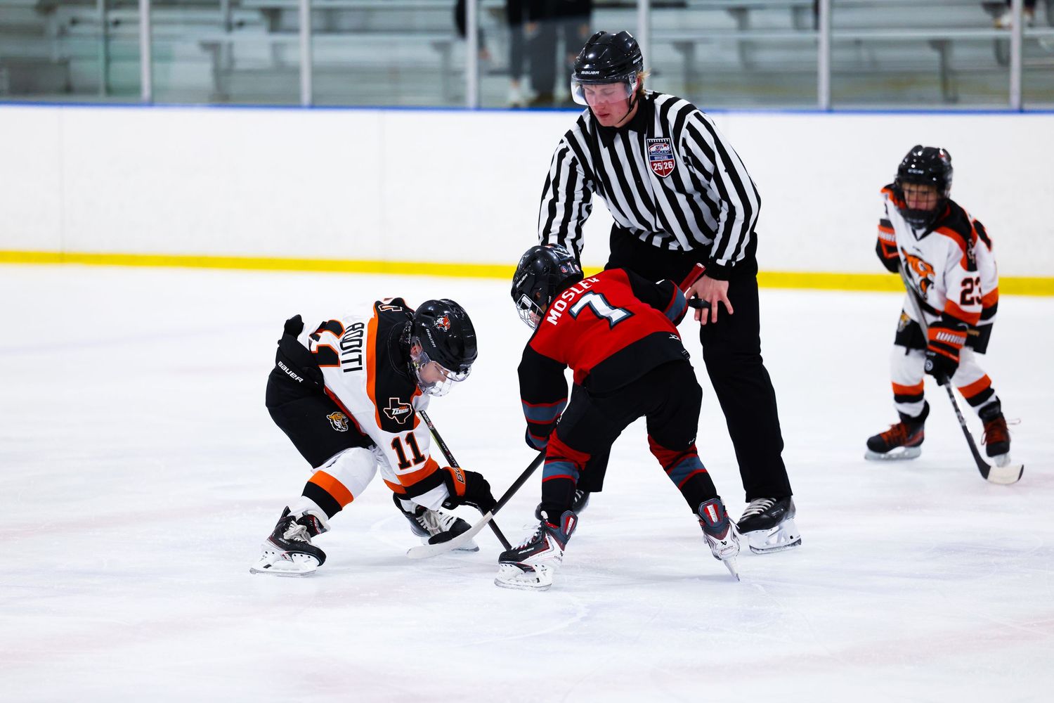 Hockey referee oversees a face-off between two youth players in black and red uniforms on indoor ice rink.