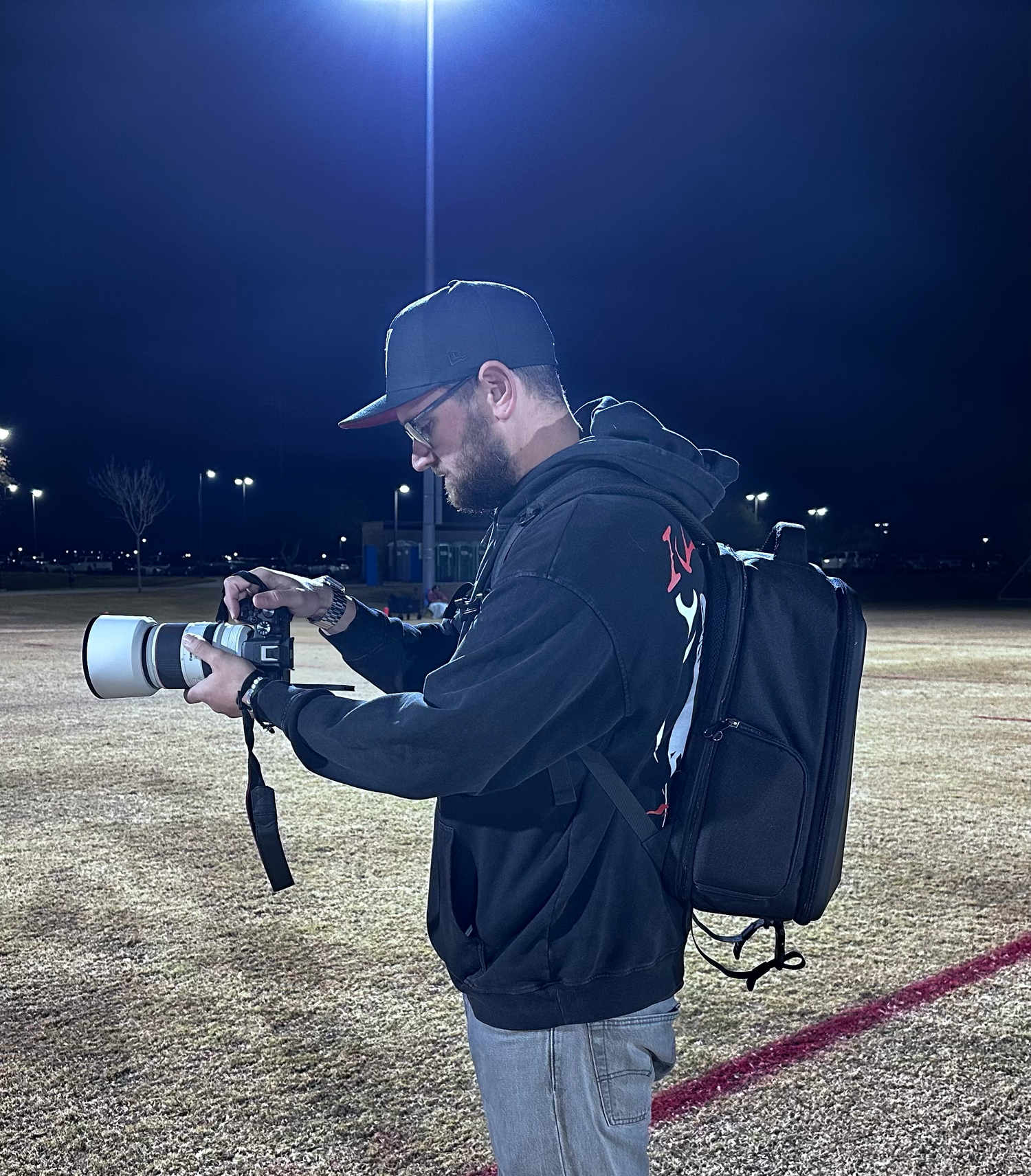 Man with backpack using a professional camera with telephoto lens on a sports field at night under bright stadium lights.