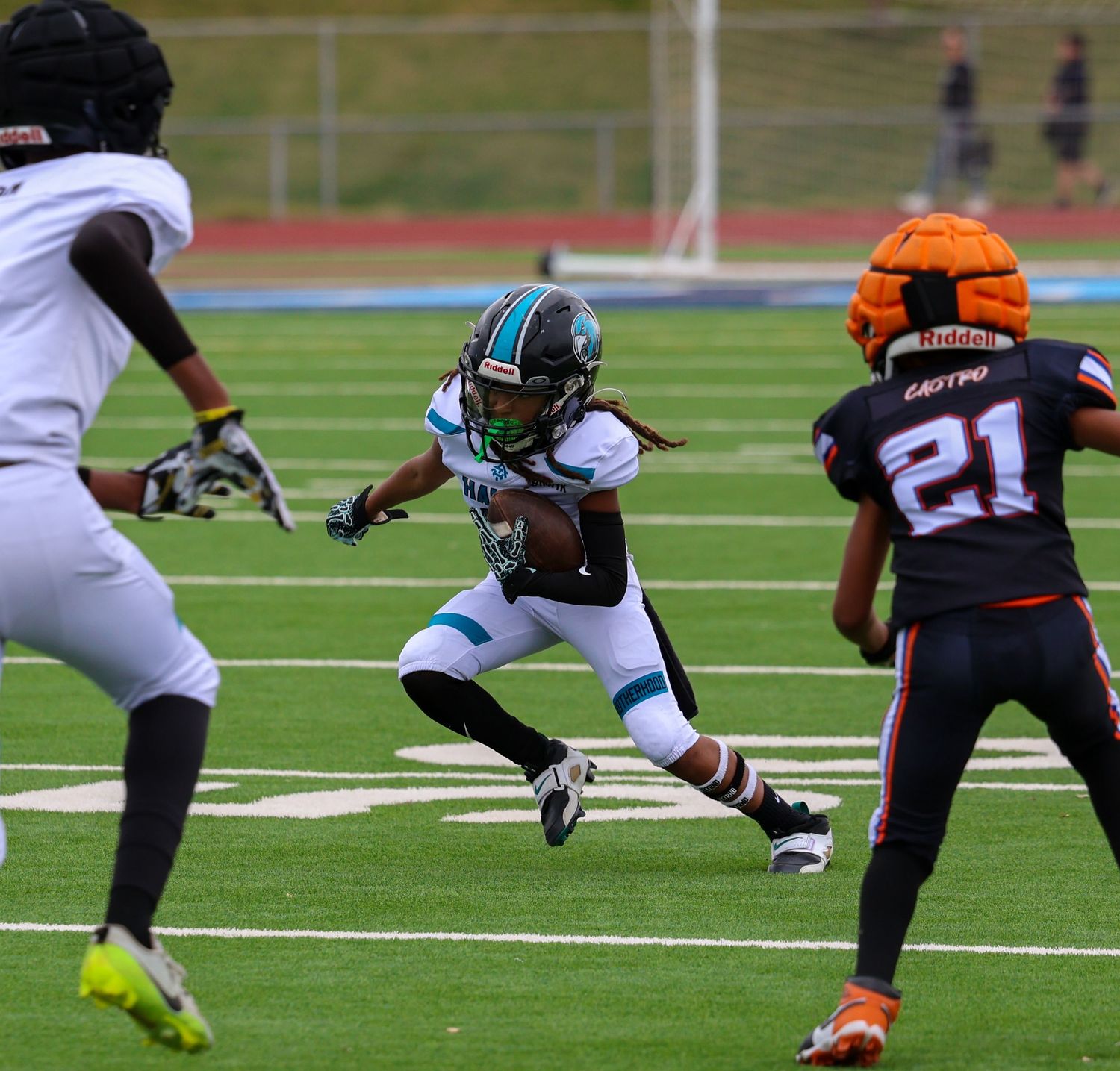 A football player in teal uniform runs with the ball between two defenders during a competitive game.