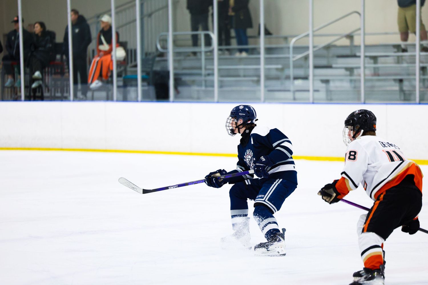 Two ice hockey players compete on the rink, one in navy blue and one in orange and white uniforms.