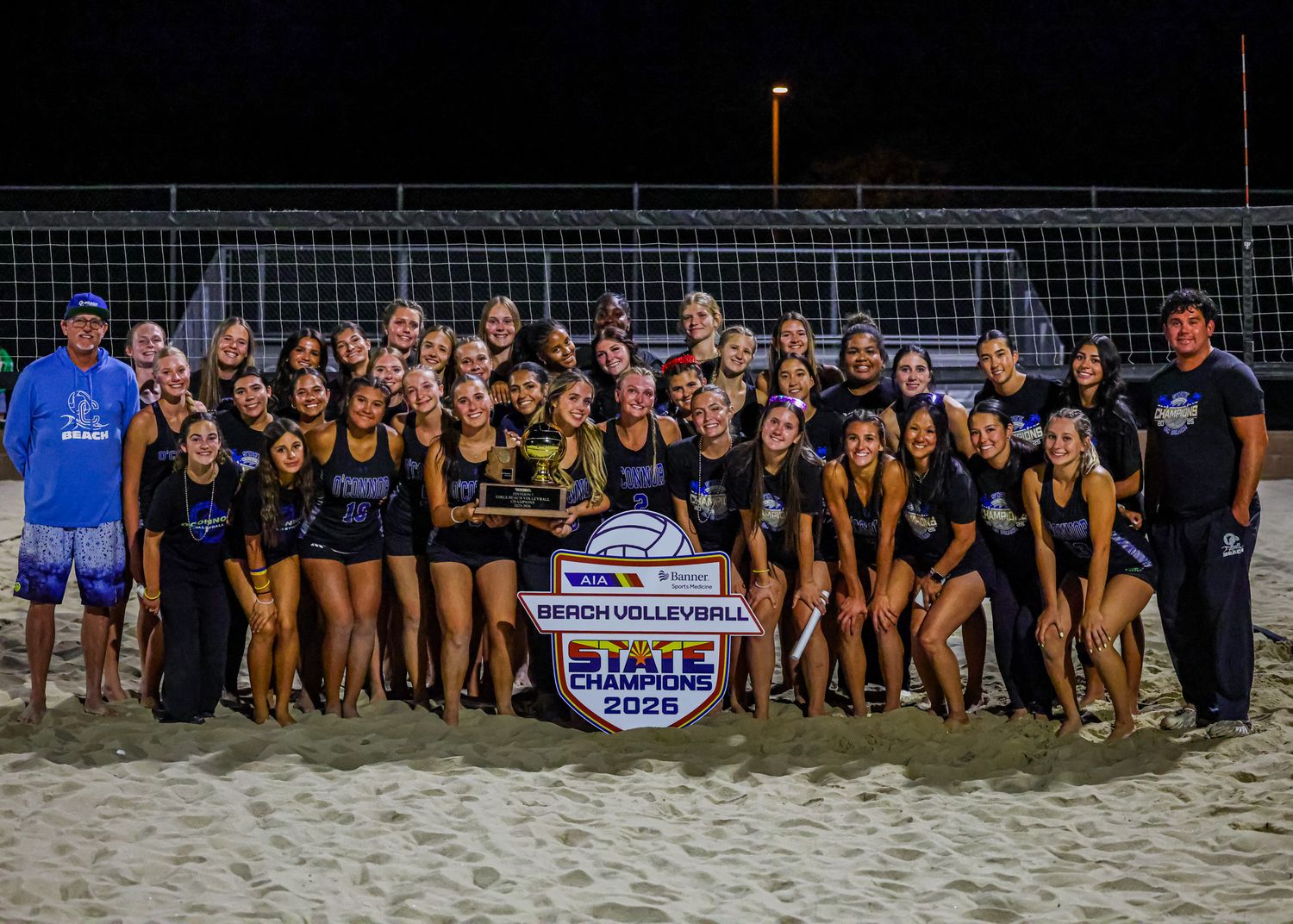 Beach volleyball team celebrates state championship win in 2025, posing with trophy on sand court at night.