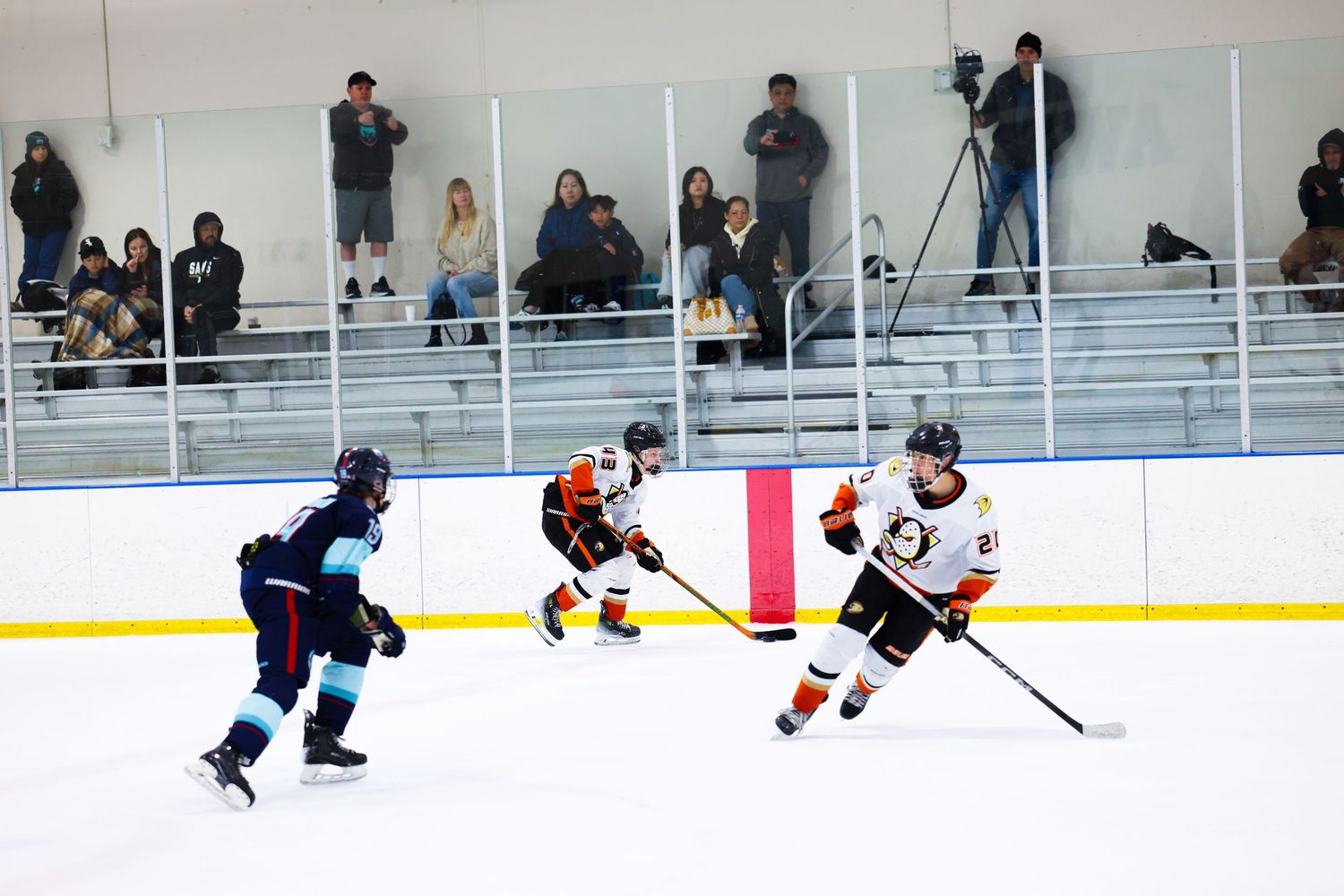 Two orange and white uniformed hockey players advance the puck against a teal-jersied defender.