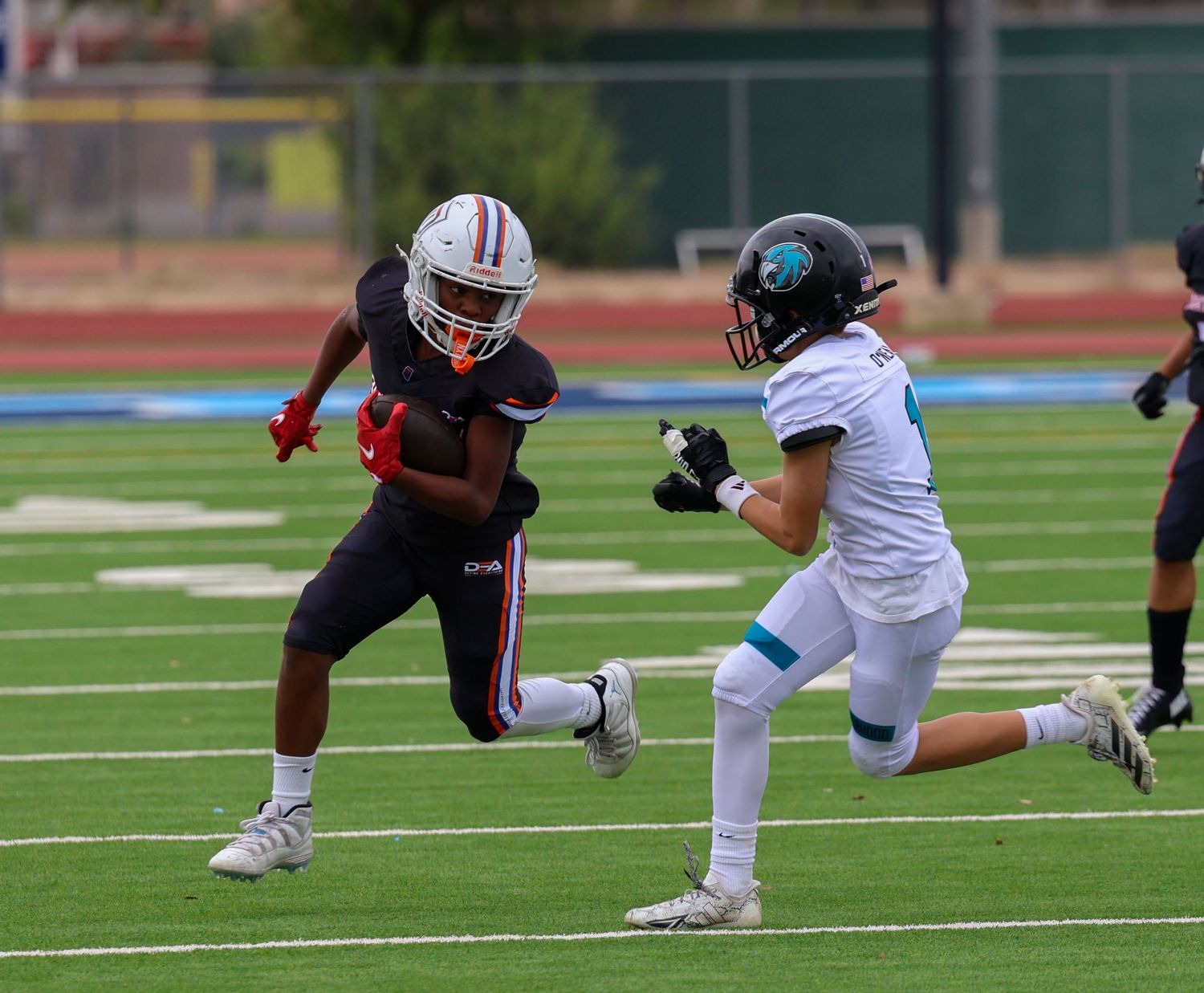 A young football player in red uniform stiff-arms a defender in teal during a high school game.