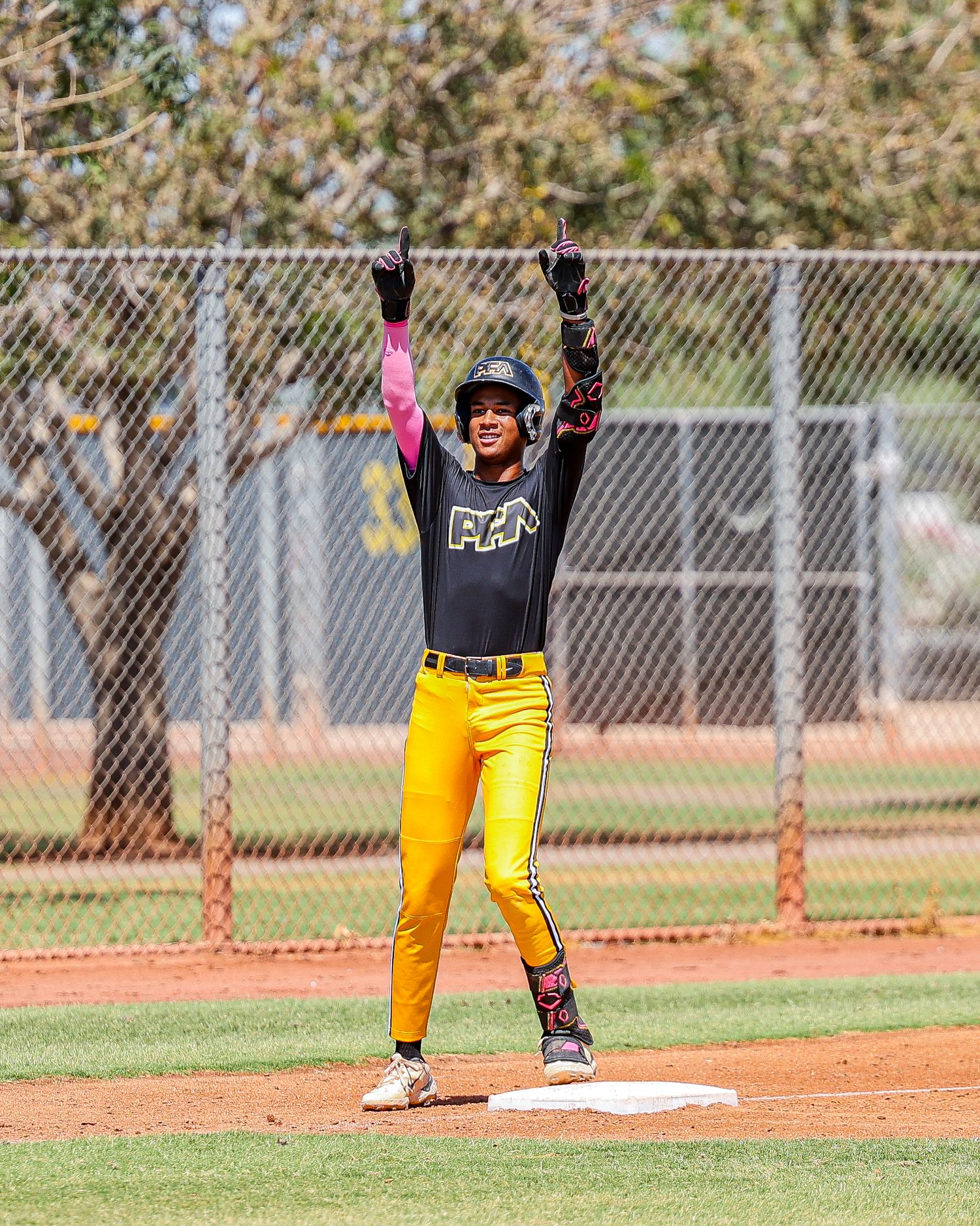 Baseball player in black PFN jersey celebrates with arms raised at base.