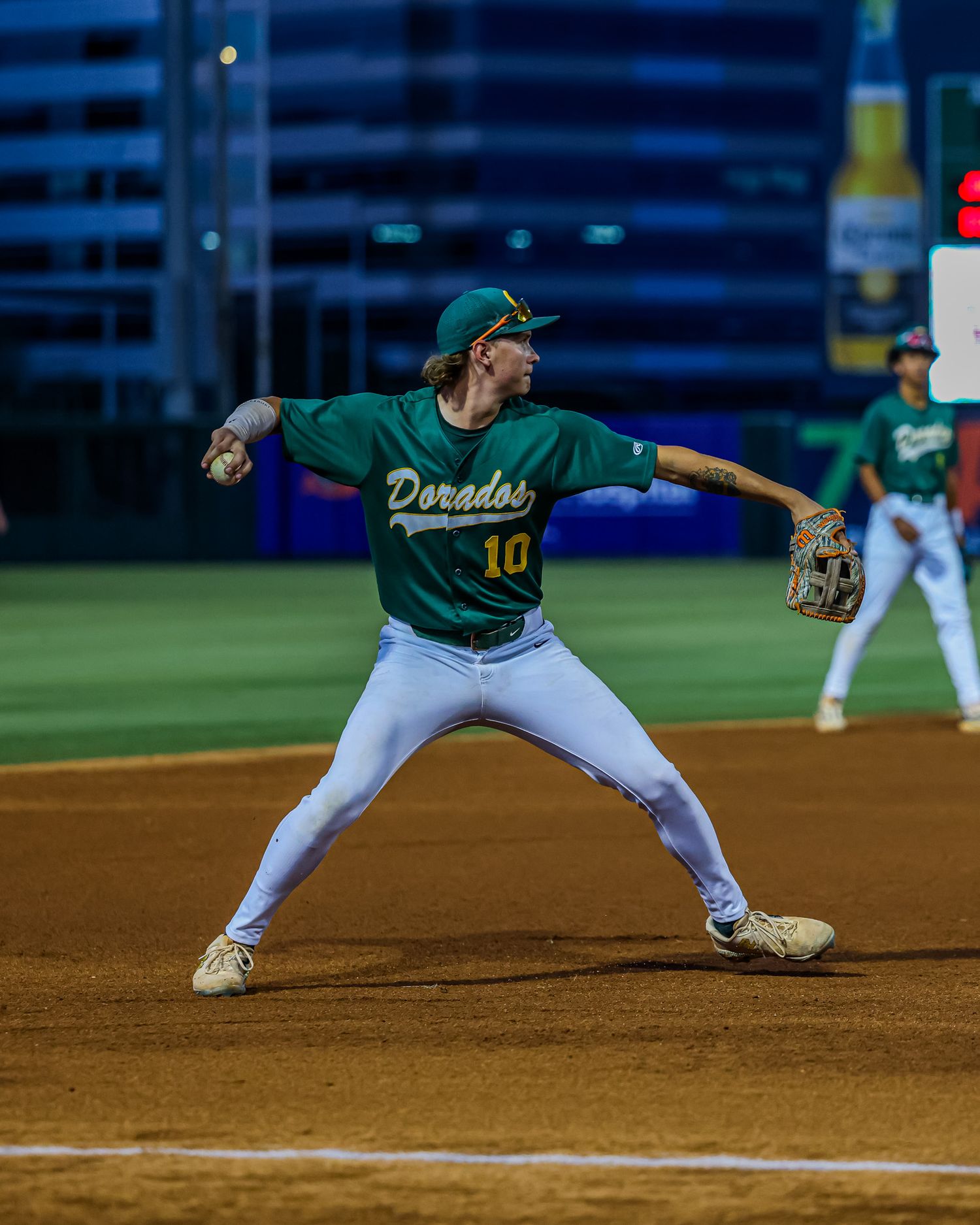 Dorados player number 10 in green uniform winds up to throw ball on baseball field.