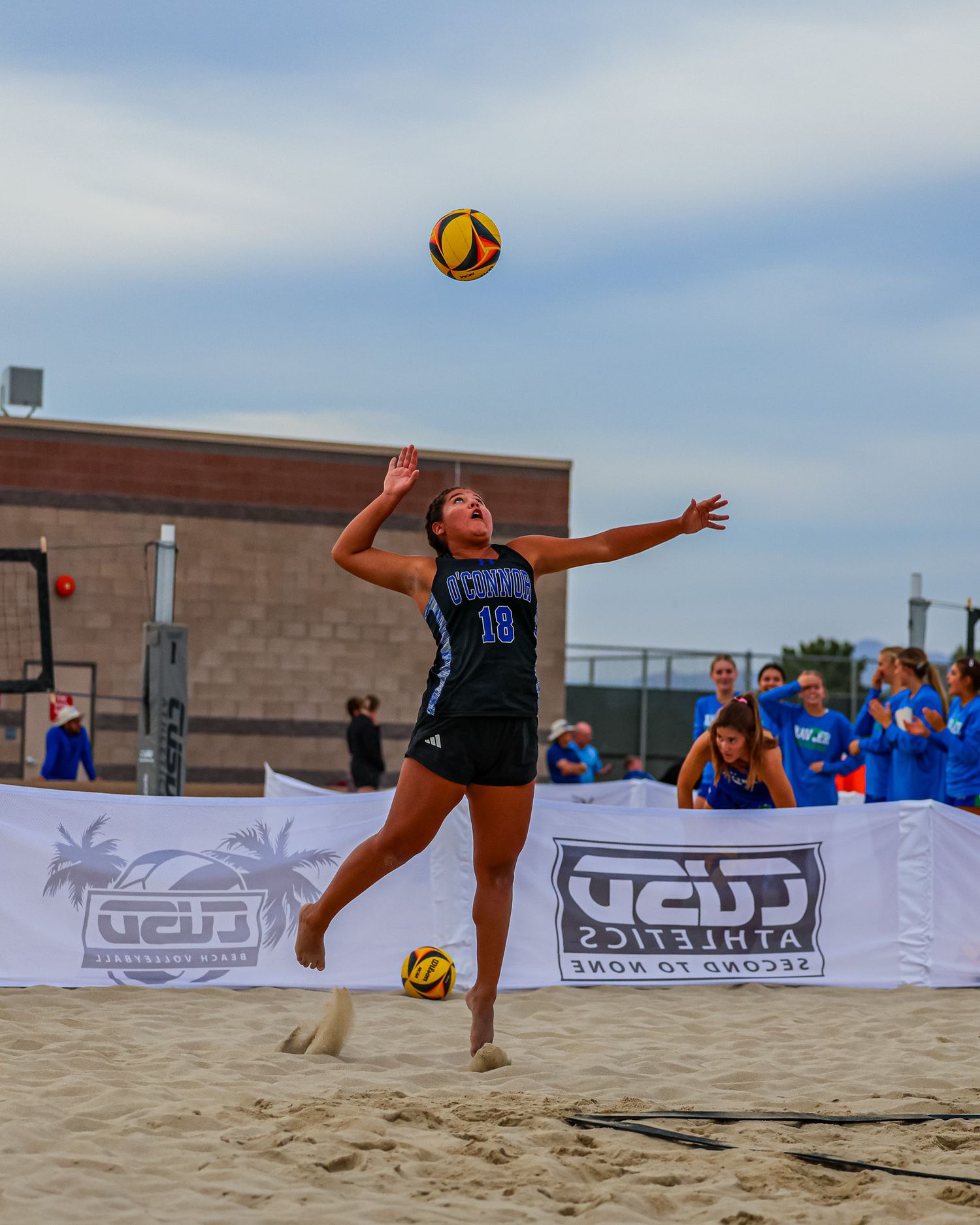 Female beach volleyball player in blue uniform jumping to serve a yellow ball at CUSD Athletics tournament.