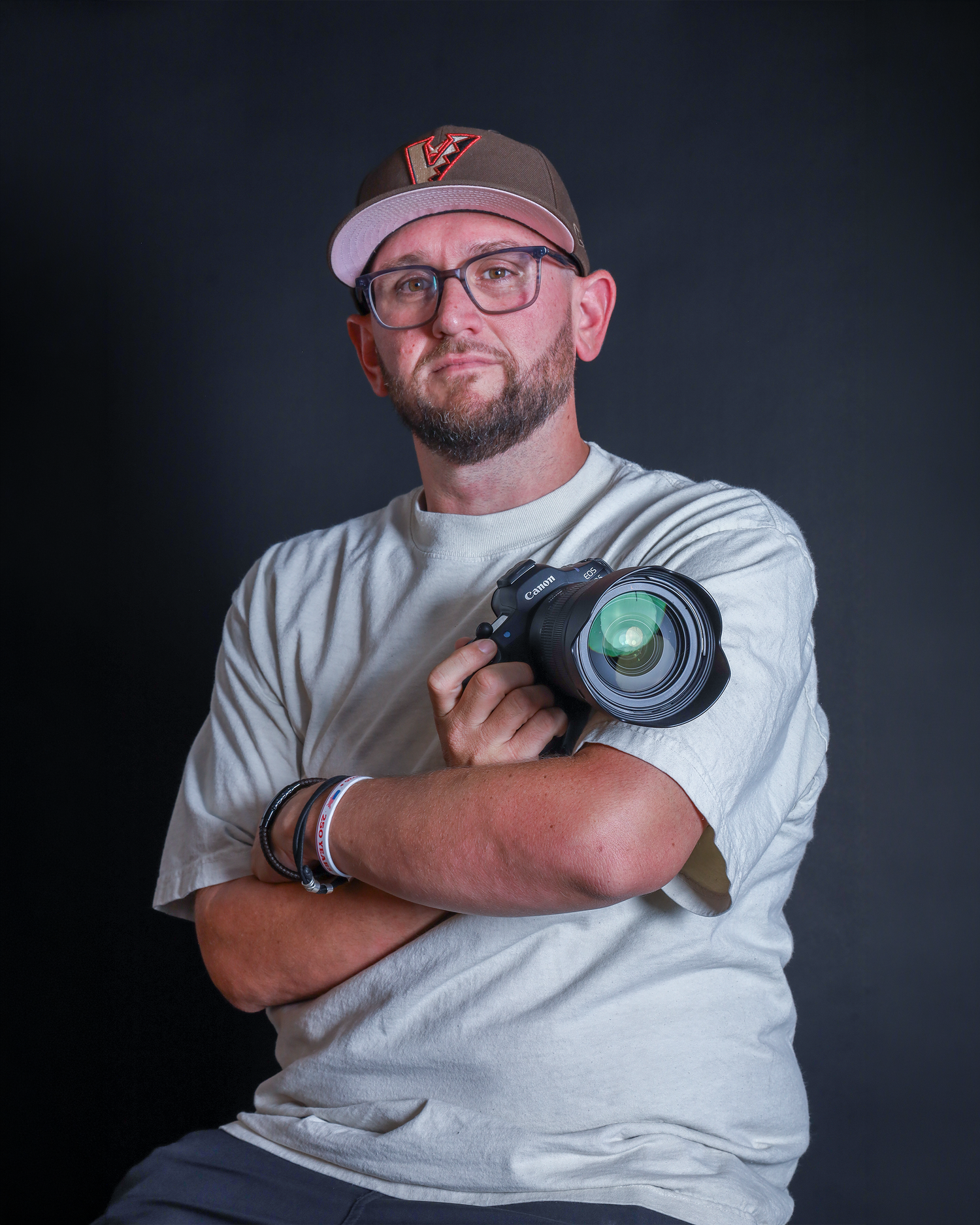 Professional photographer poses with camera and crossed arms against dark studio background.