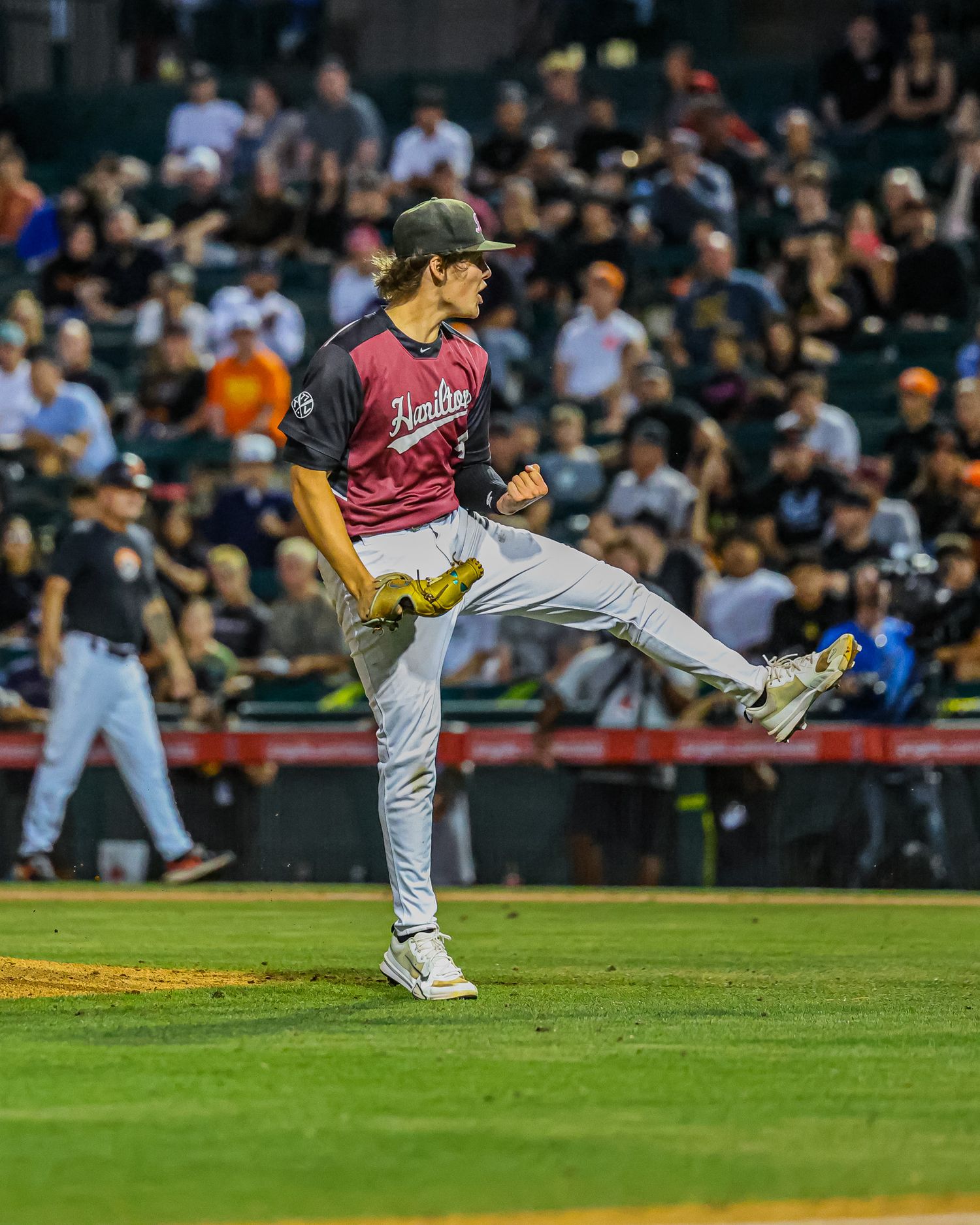 Baseball pitcher in maroon uniform celebrates with a high kick on the mound during a night game.