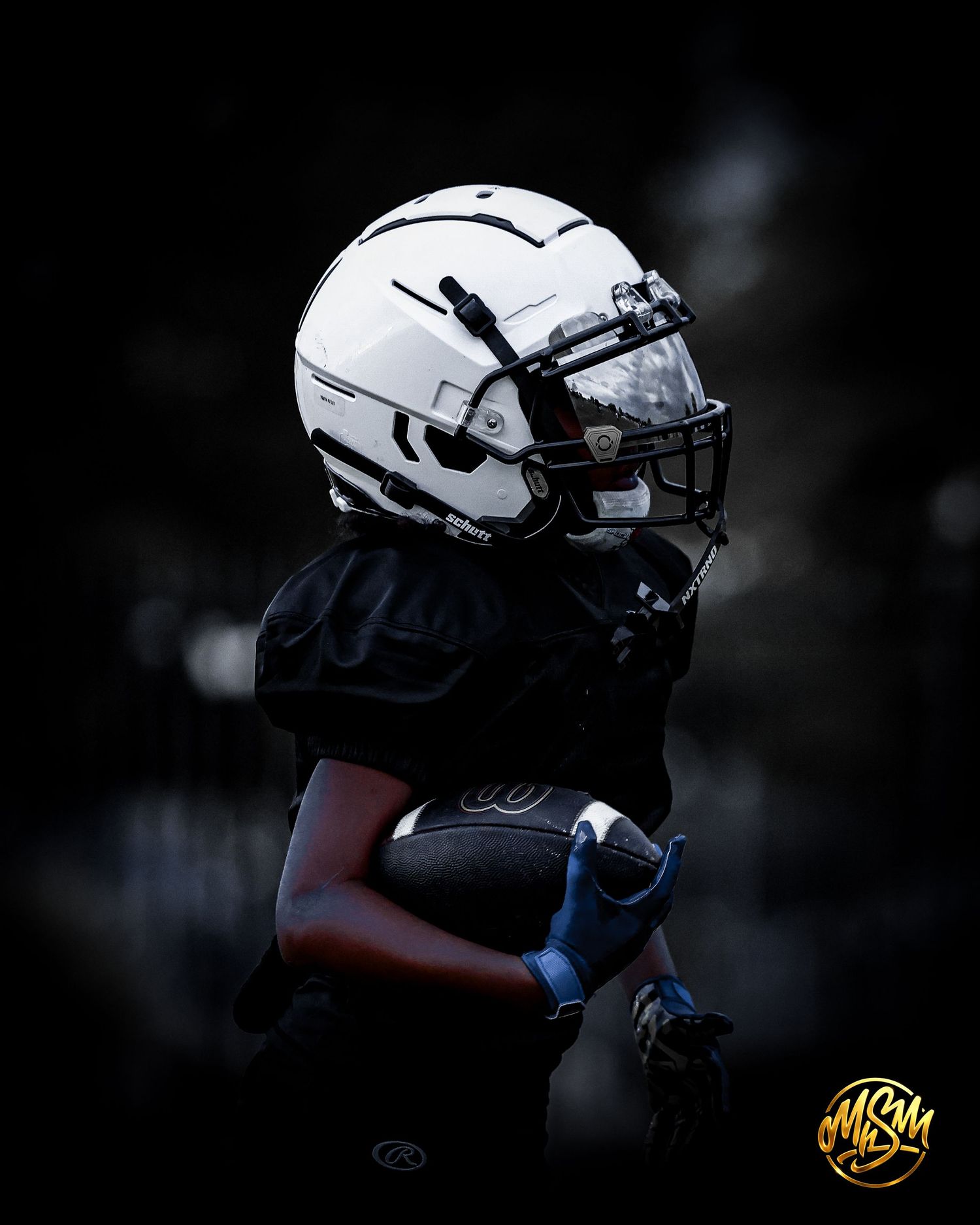 Football player in black jersey and white helmet holds ball in dramatic dark lighting.