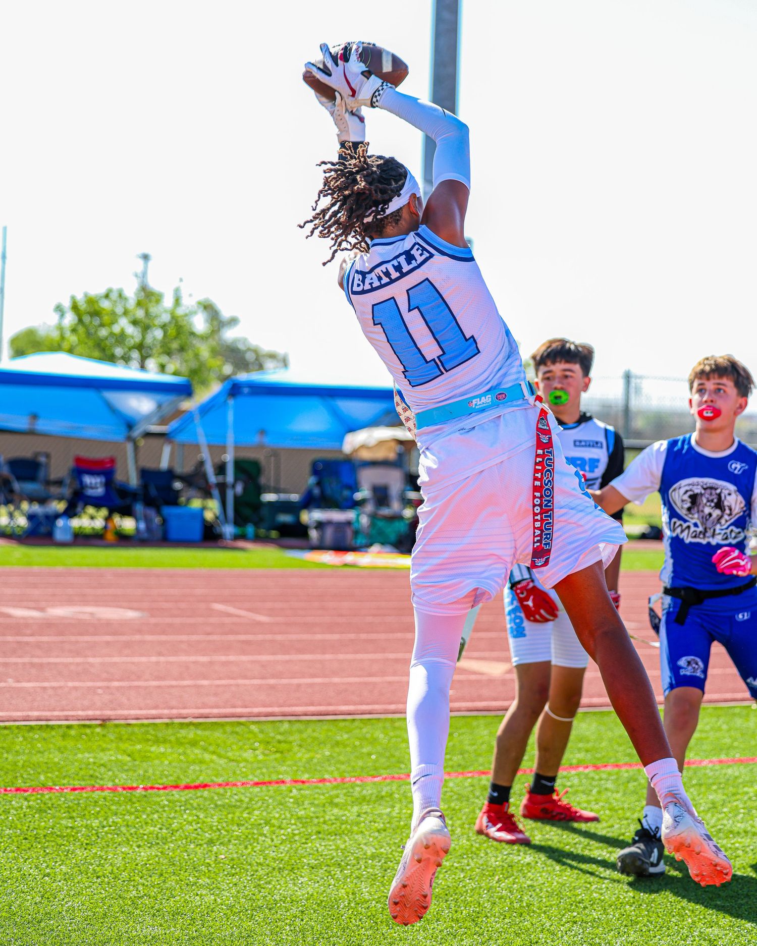 Youth flag football player #11 in white leaps to catch pass during outdoor game.