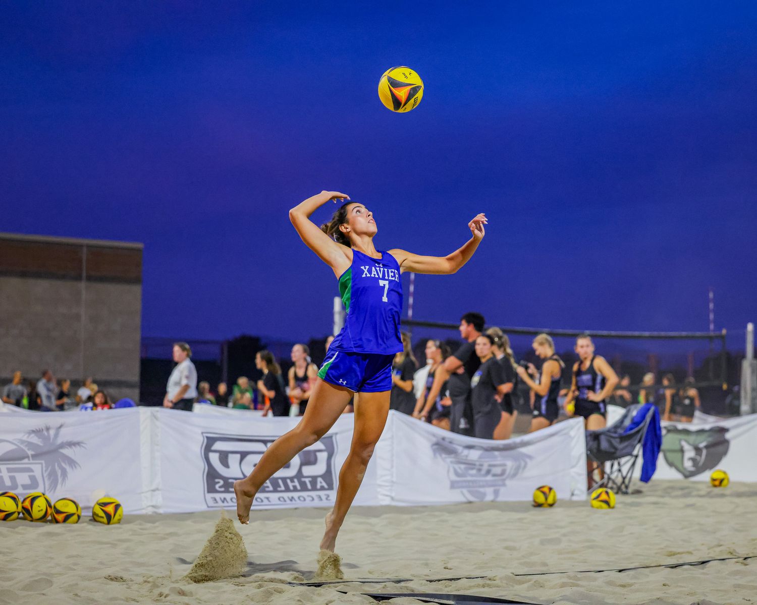 Female beach volleyball player in blue jersey number 7 serving the ball under a twilight sky.