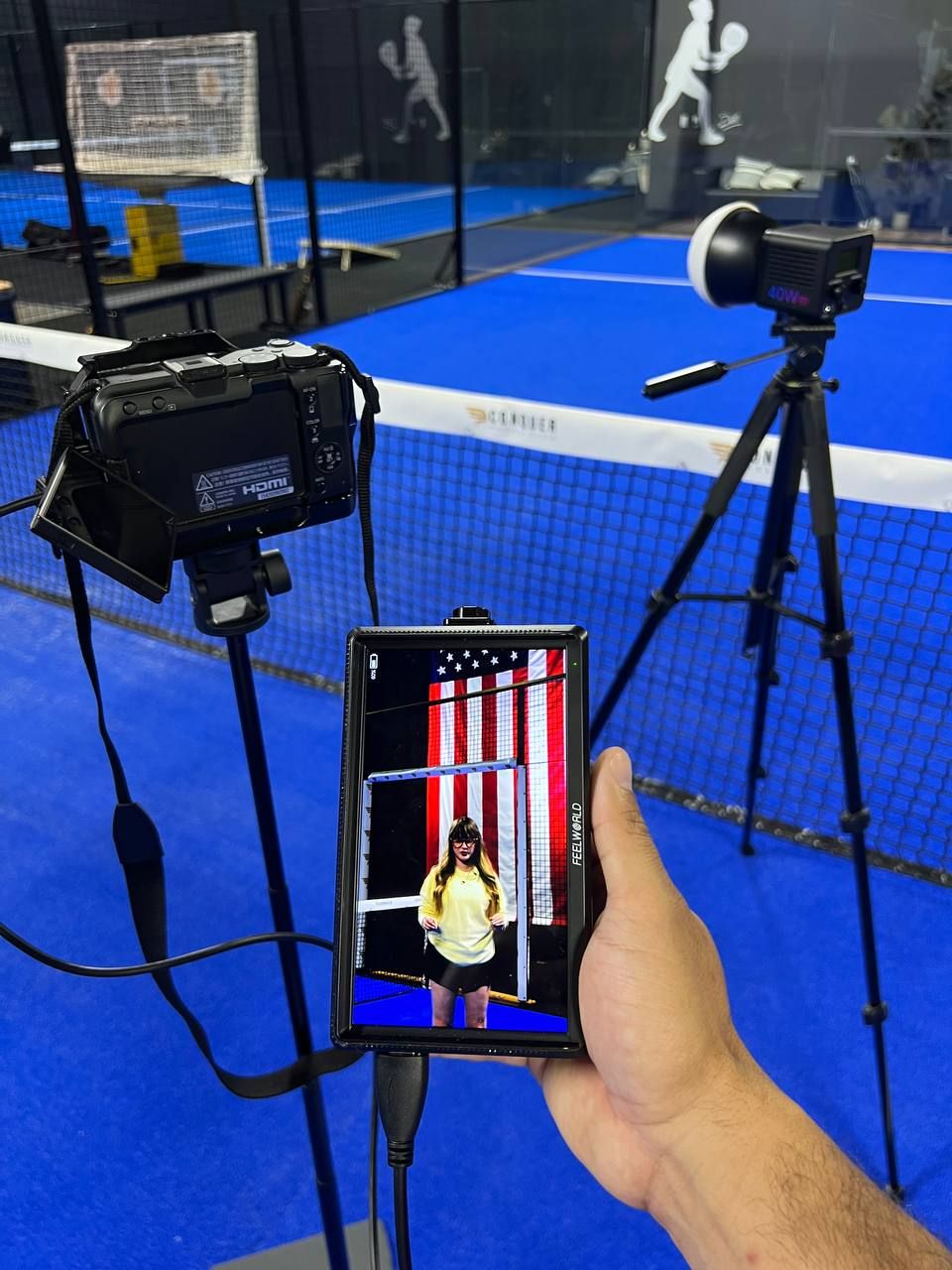 Hand holding a smartphone displaying a woman posing, with padel court cameras on tripods in background.