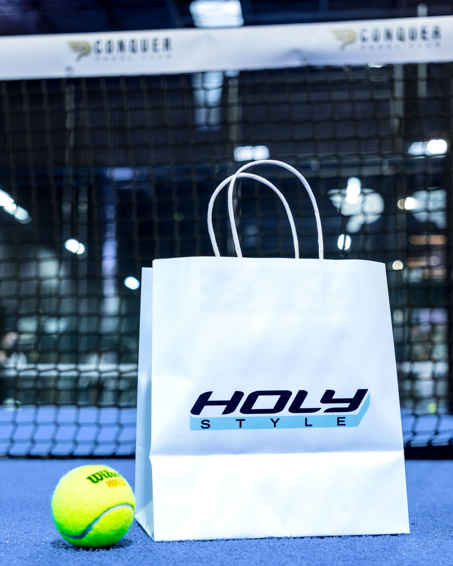 White Holy Style branded gift bag next to a yellow Wilson tennis ball on a blue padel court.