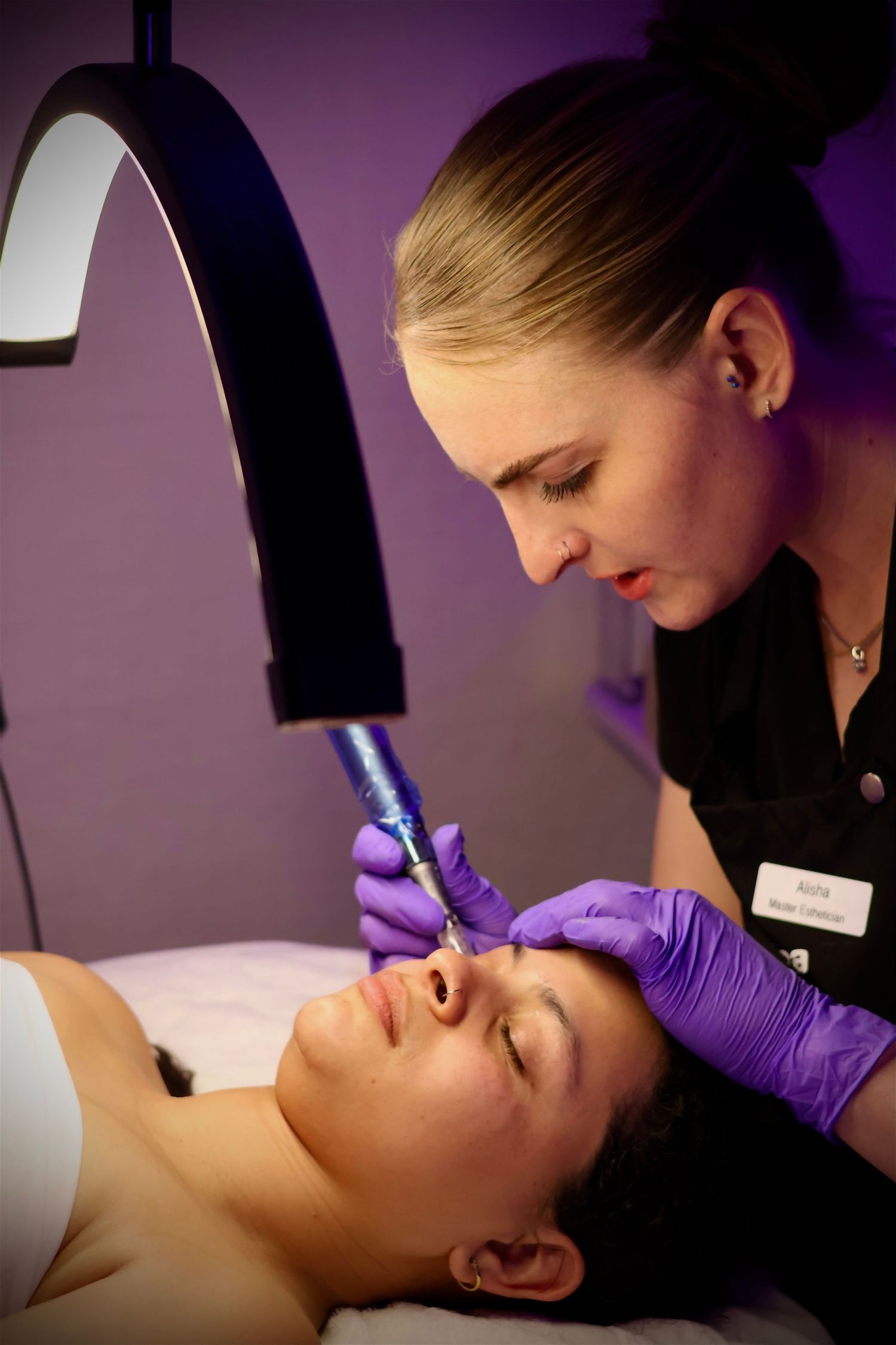 Blonde esthetician using a microneedling device on a female client under a professional treatment lamp.