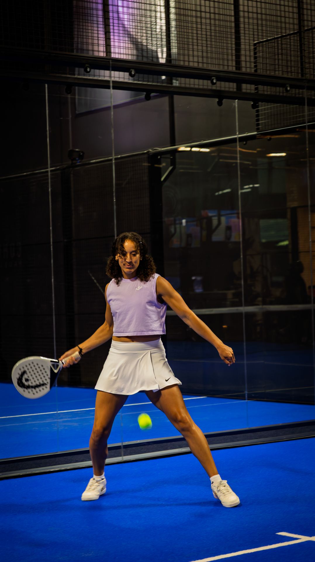 Woman in white skirt and purple top playing padel tennis on a blue indoor court, mid-swing at a yellow ball.