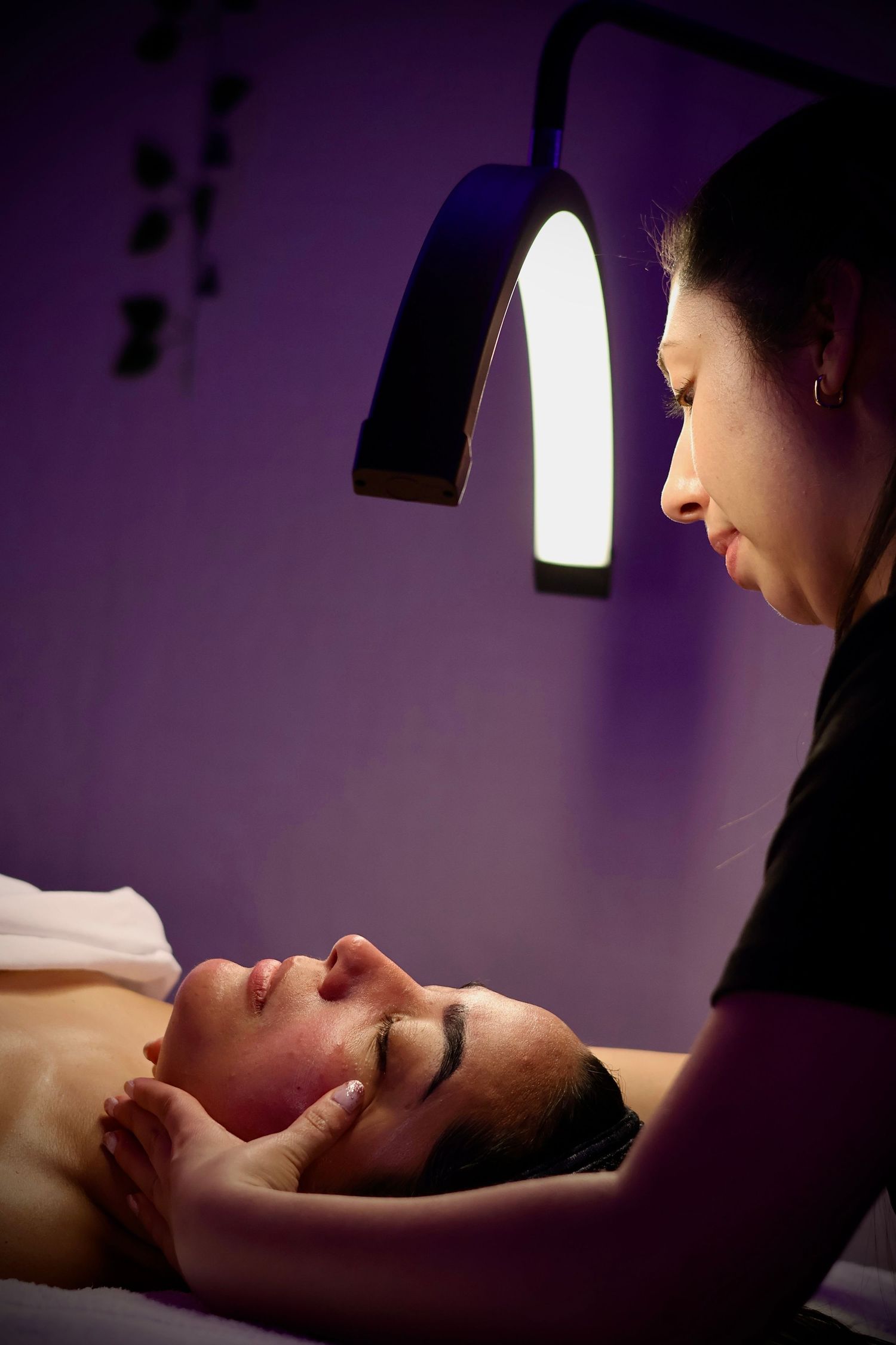 Esthetician examining a female client lying on a treatment bed under a curved salon lamp.