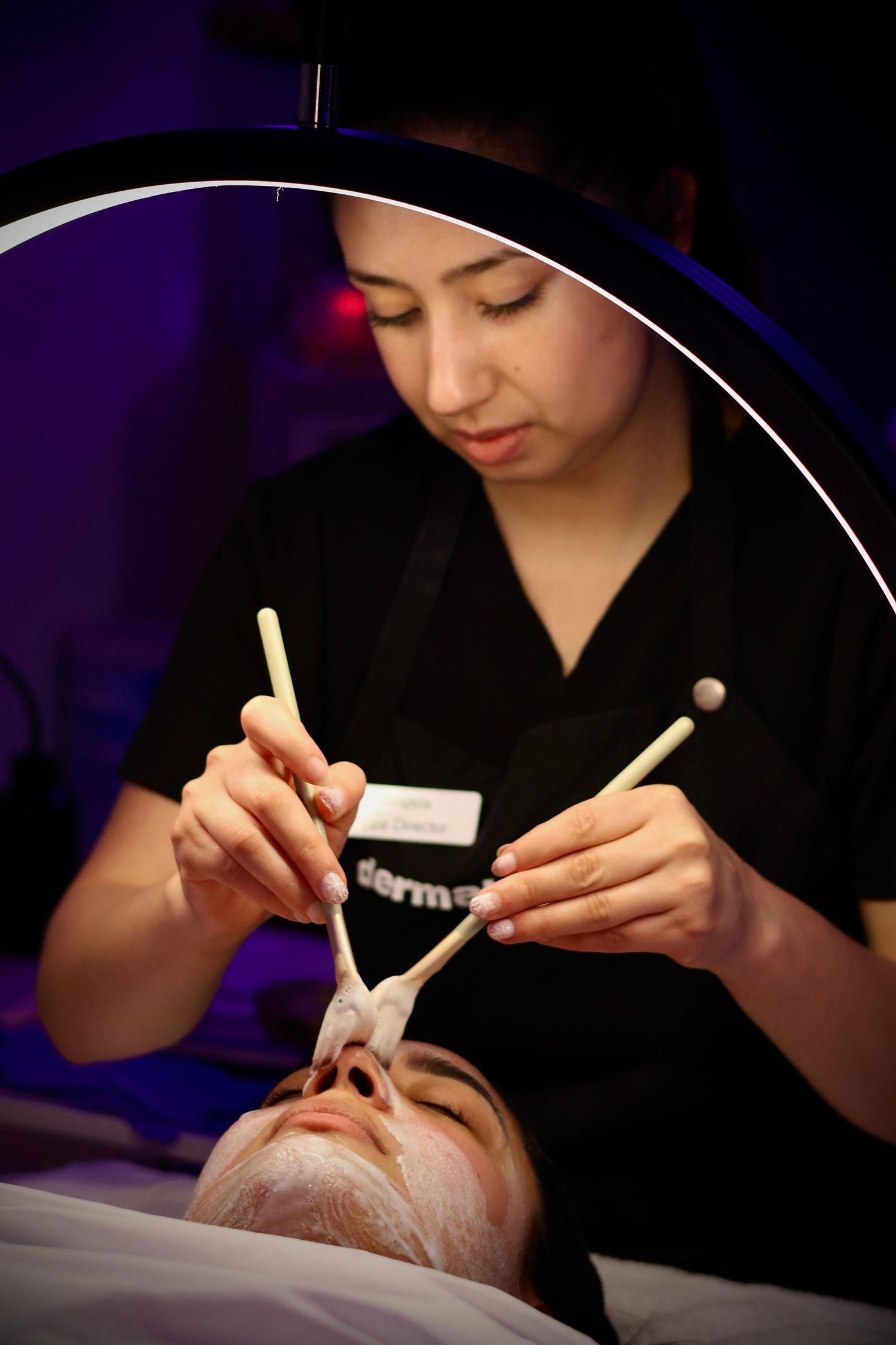 Esthetician applying a white facial mask with brushes to a client under a circular treatment lamp.