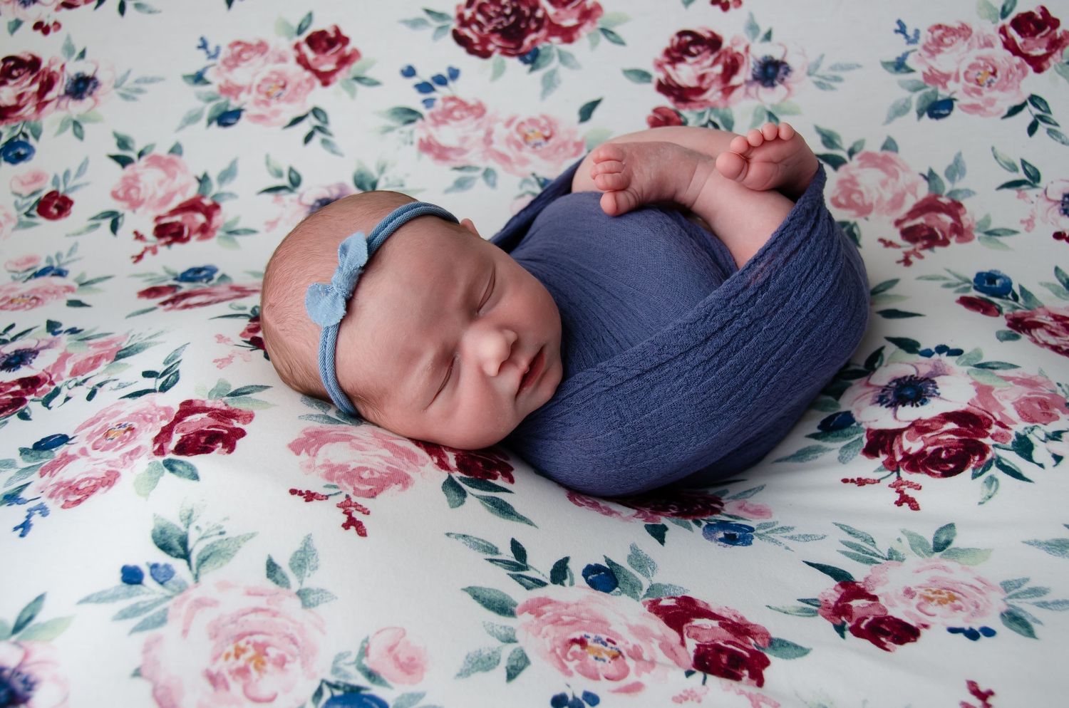 newborn girl with blue head bow with matching blue blanket laying on blanket with flowers in professional home photography studio henderson