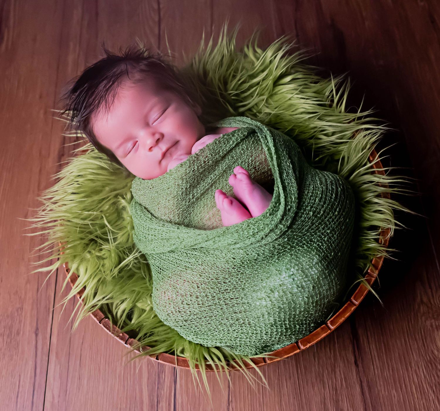 professional newborn portrait of baby boy wrapped in green blanket laying wooden bowl with green fur