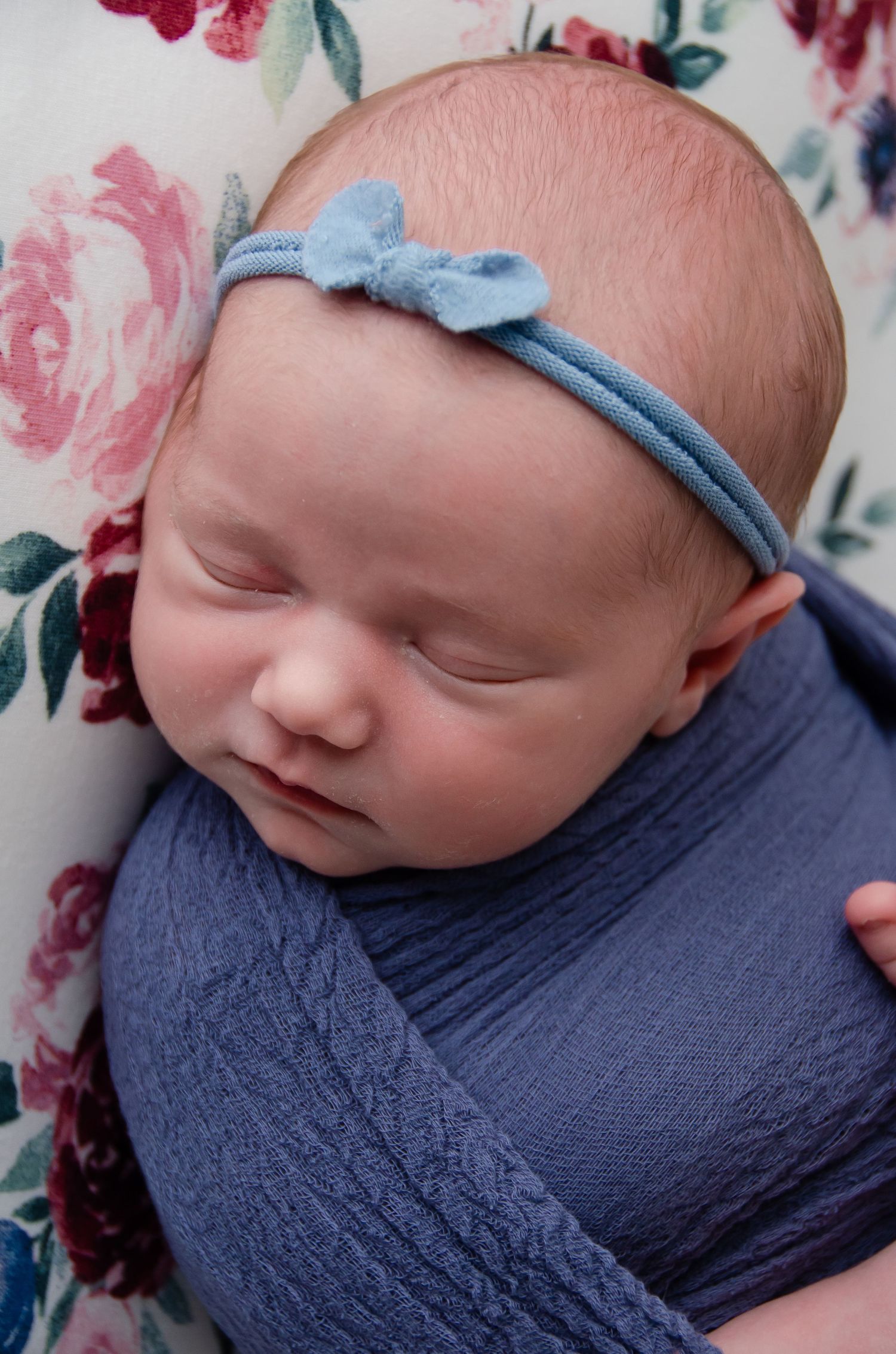 newborn girl with blue head bow with matching blue blanket laying on blanket with flowers in professional home photography studio