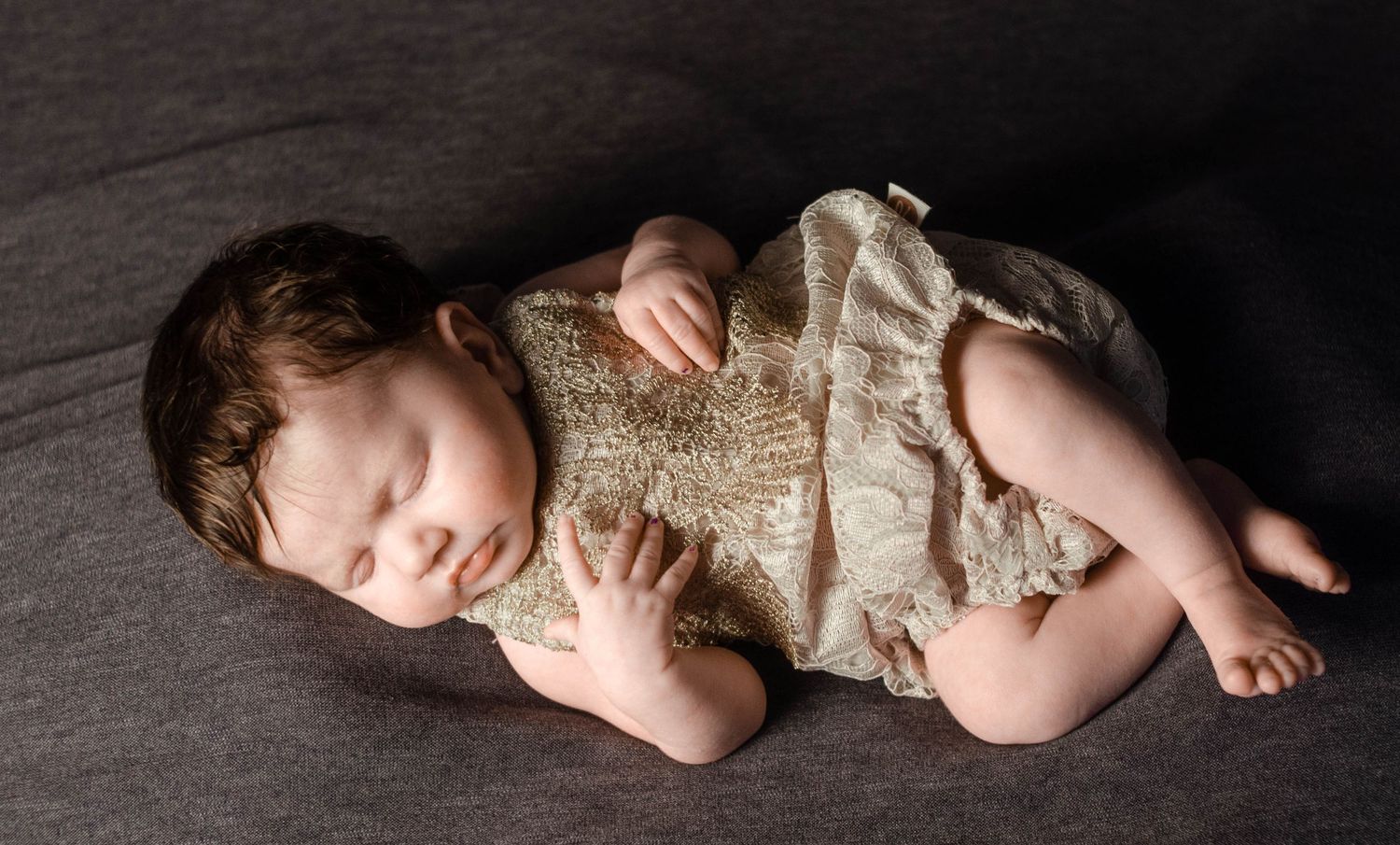 baby girl in tan lace outfit laying on grey blanket in newborn photography home studio