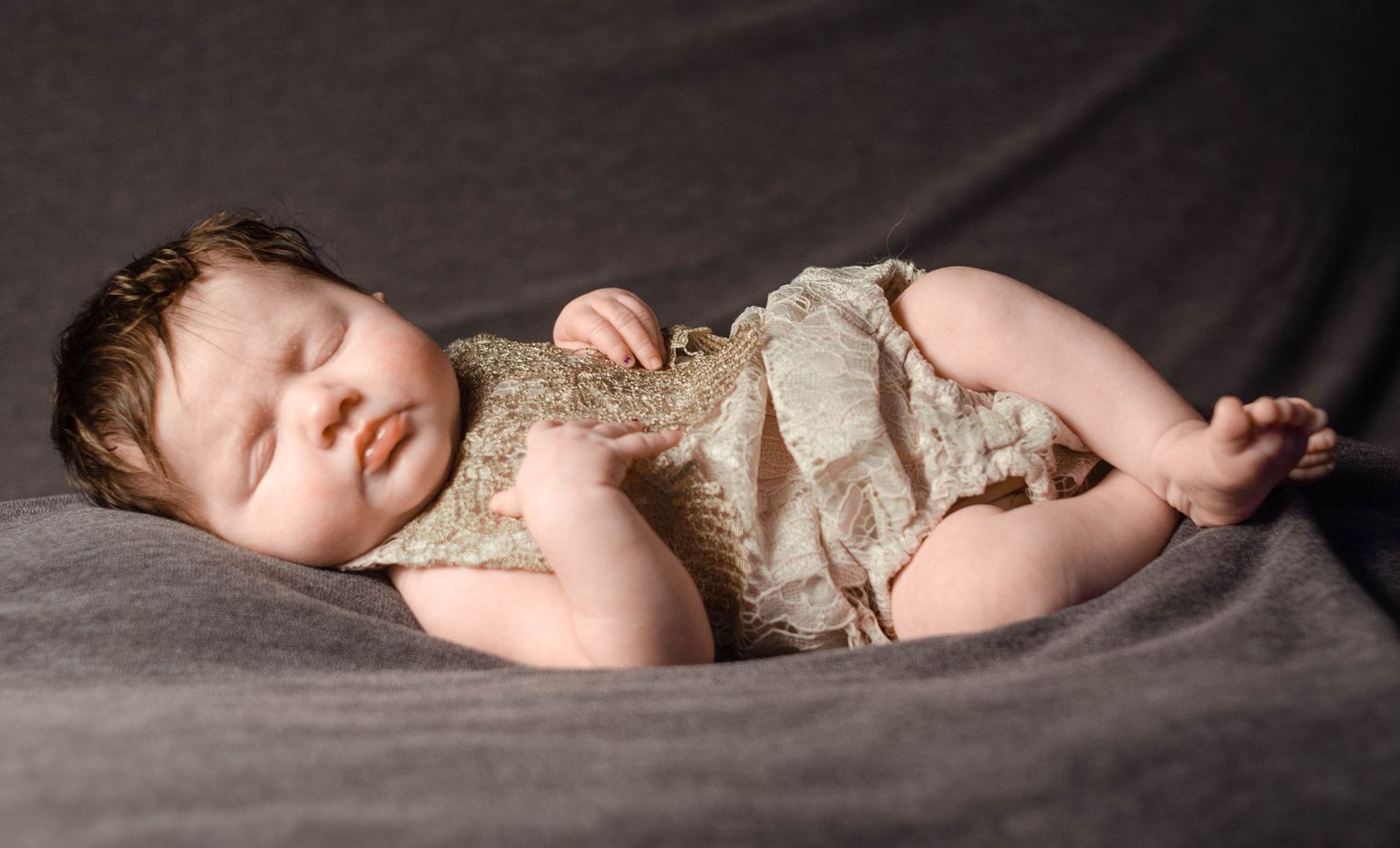 baby girl in tan lace outfit laying on grey blanket in newborn photography home studio