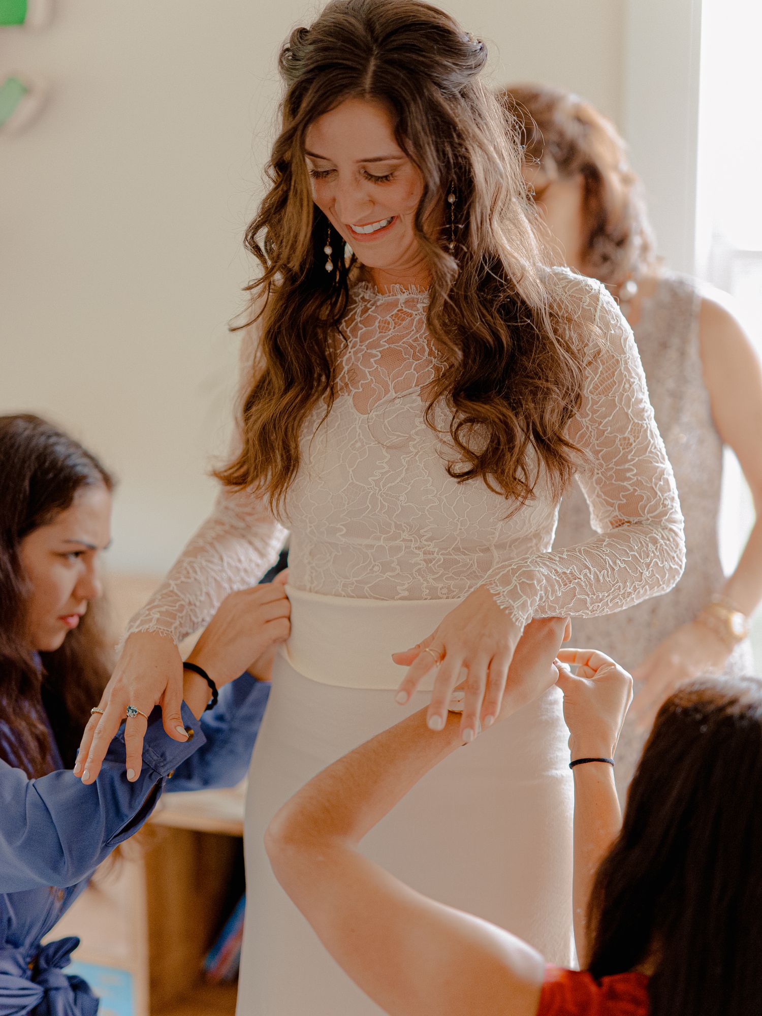 Bride in white lace long-sleeve wedding gown smiling as bridesmaids help her prepare for her wedding day.