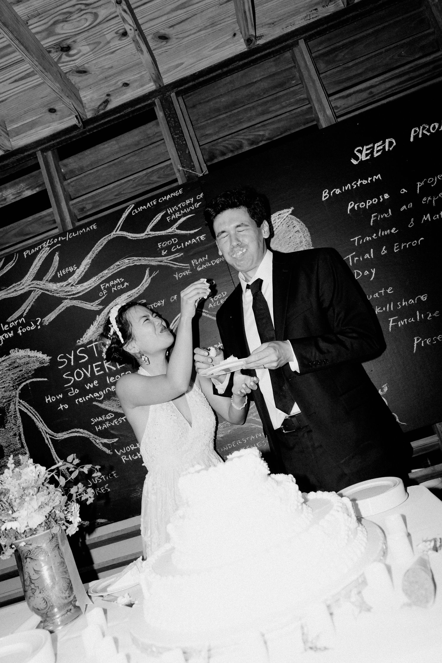 Black and white photo of bride and groom laughing while cutting their wedding cake at reception.