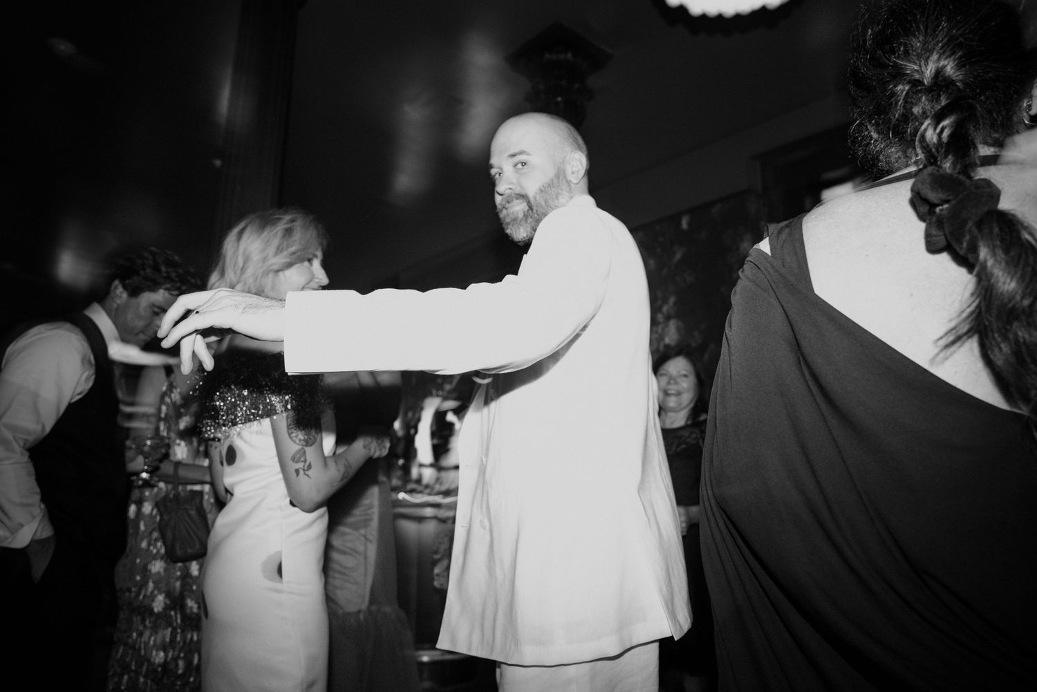 Black and white photo of bearded man in white suit dancing with woman at reception.