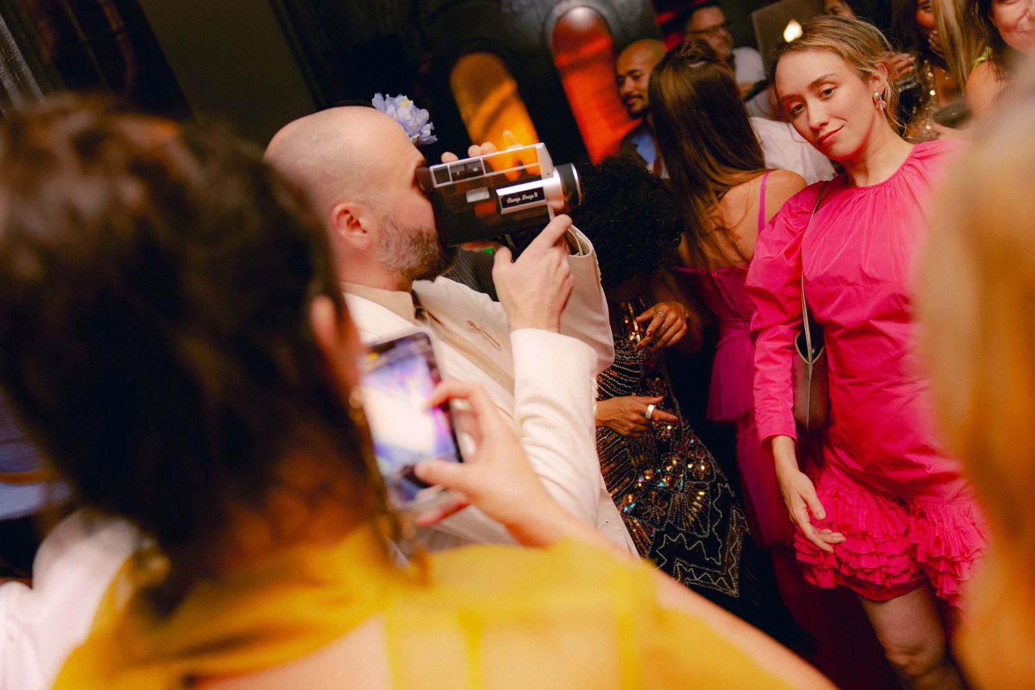 Bald groom photographs a guest in a pink dress at a lively, crowded wedding reception.