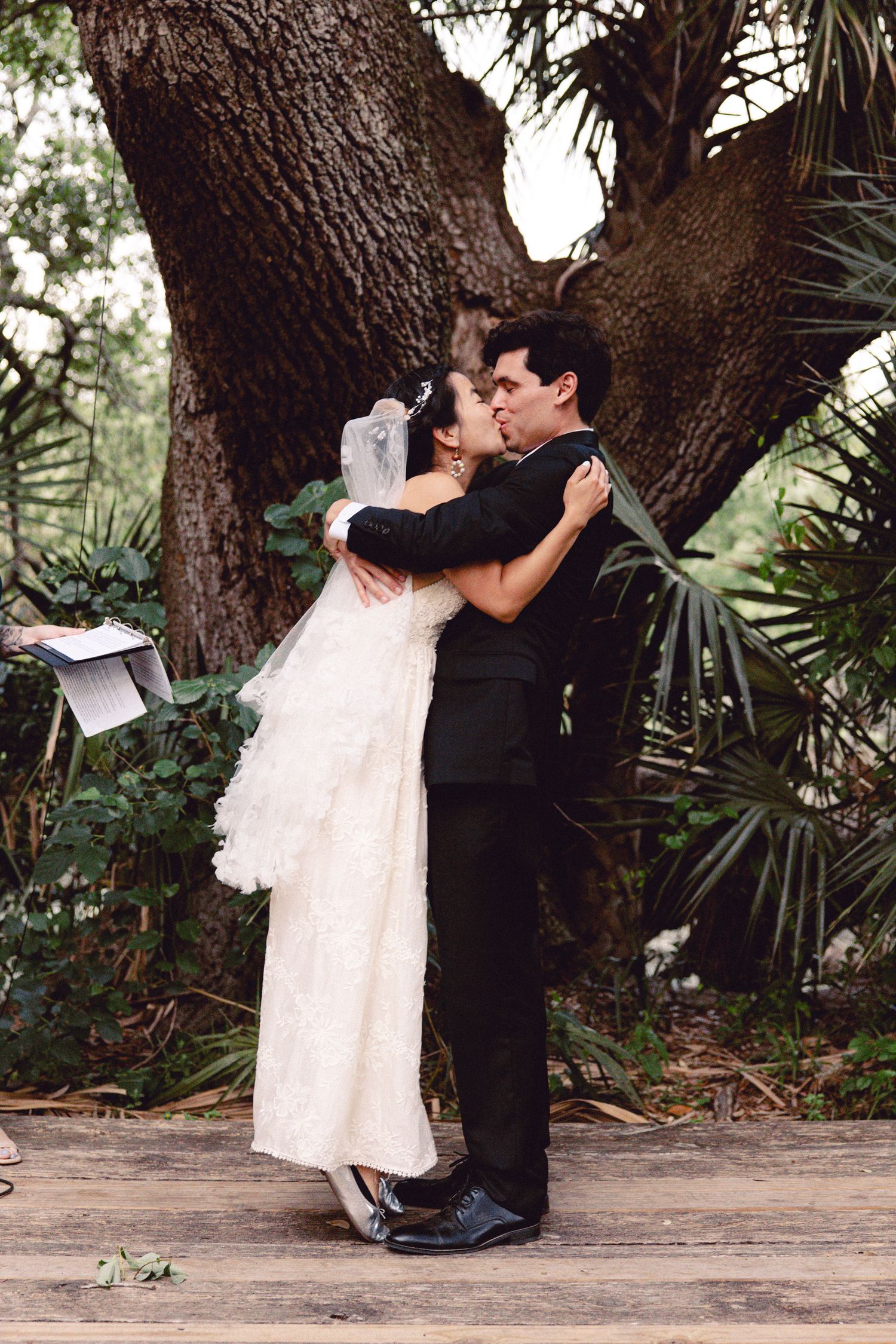 A bride and groom share their first kiss under a large tree at an outdoor woodland wedding.